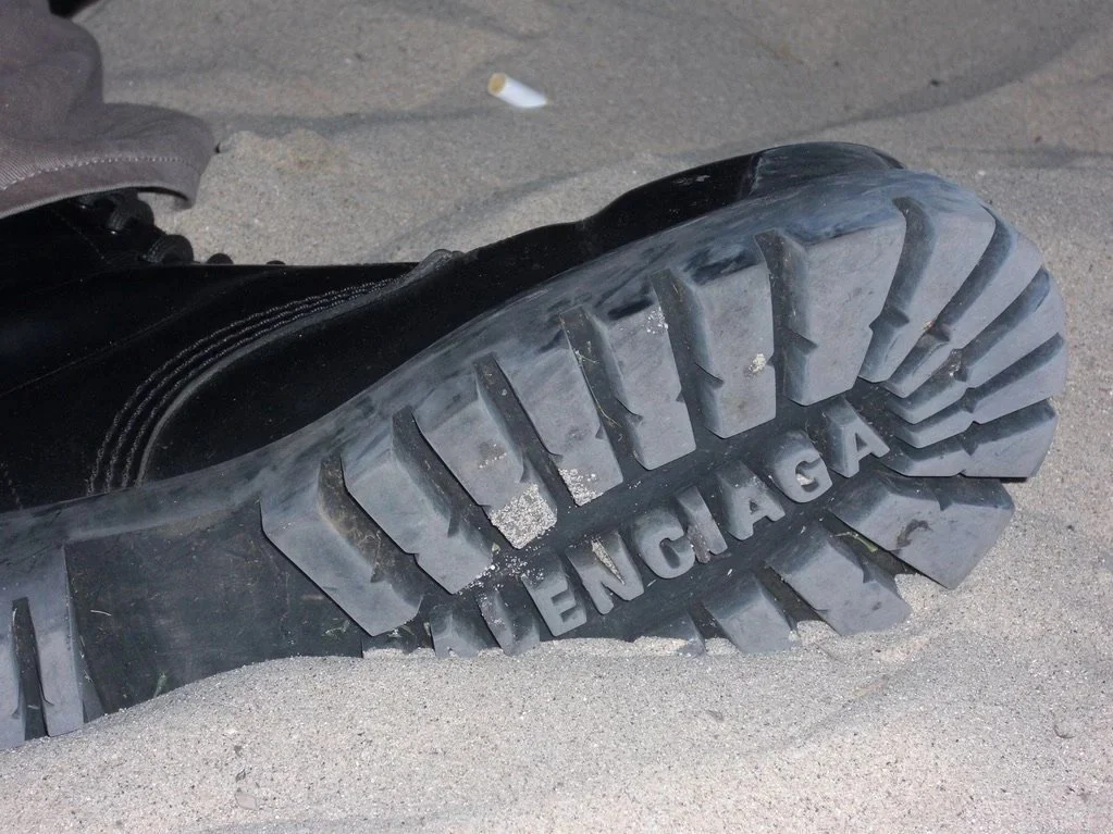 A fallen black boot with a thick tread sole on sandy ground, with the word "BALENCIAGA" embossed on the sole.