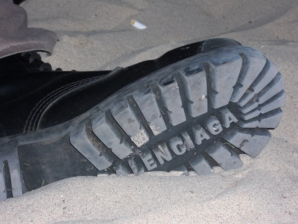 A black combat boot with a thick sole, partially buried in the sand. The sole has the word 'BALENCIAGA' embossed on it.