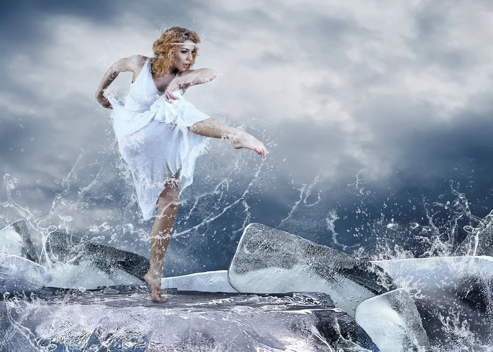 A woman in a white dress balancing on a large ice block, surrounded by splashing water, with a cloudy sky in the background.
