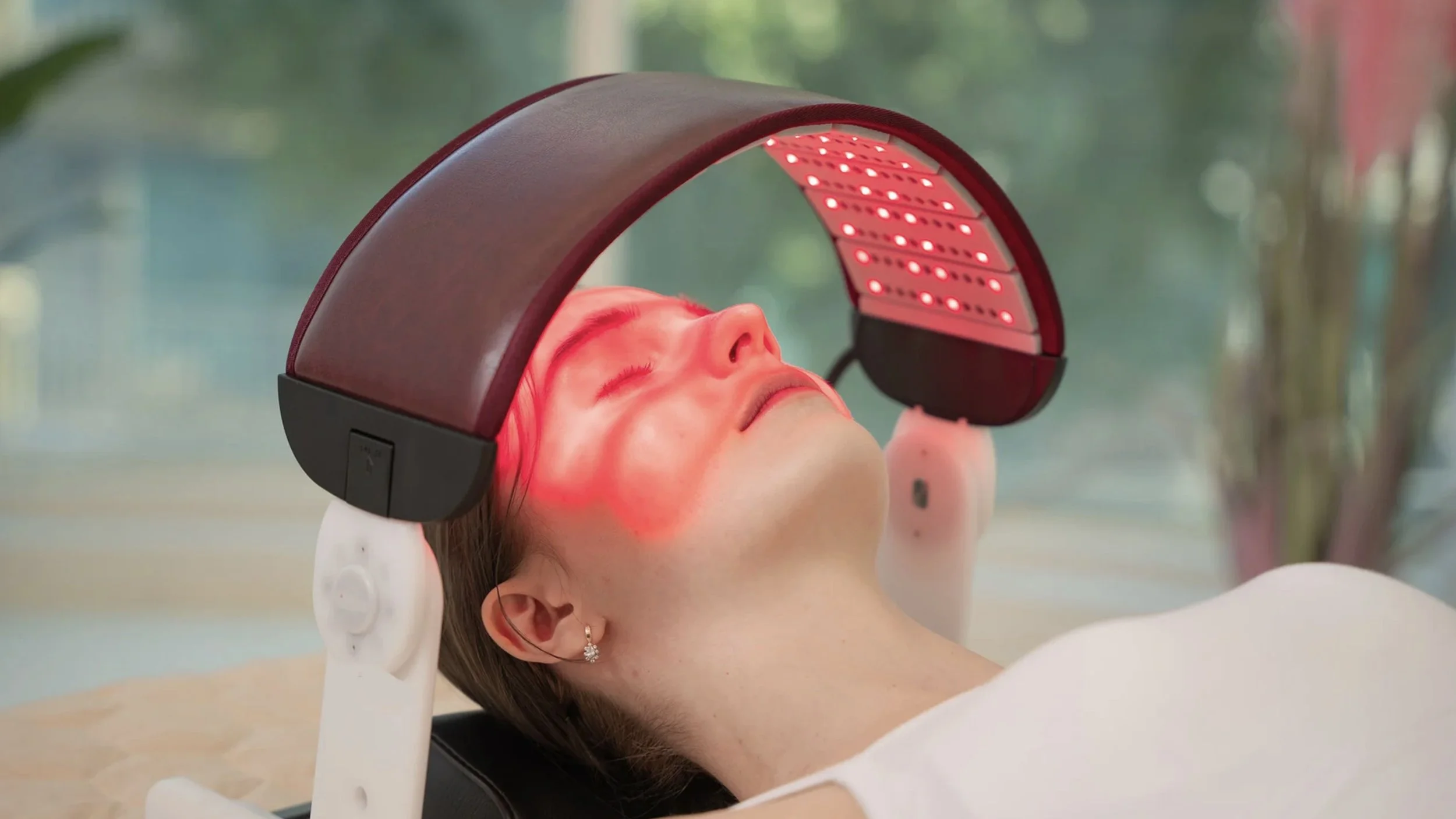 A woman receiving red light therapy on her face while lying down, with a curved red LED light device positioned over her face.