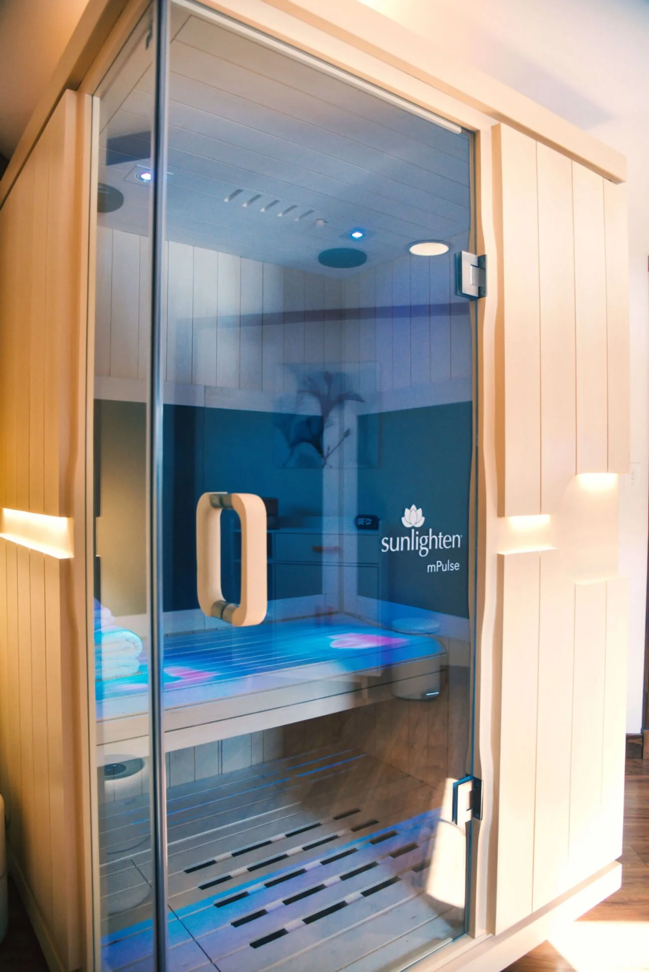 A wooden sauna room with a glass door, inside features a bench, towels, and a control panel, labeled 'Sunlighten mPulse'.