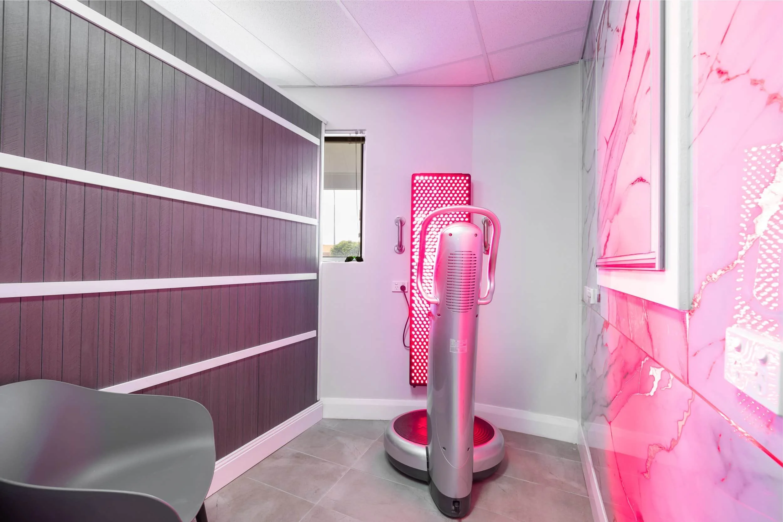 Salon room with RED/IR LED light therapy device, a gray chair, purple textured wall, and pink marble wall.