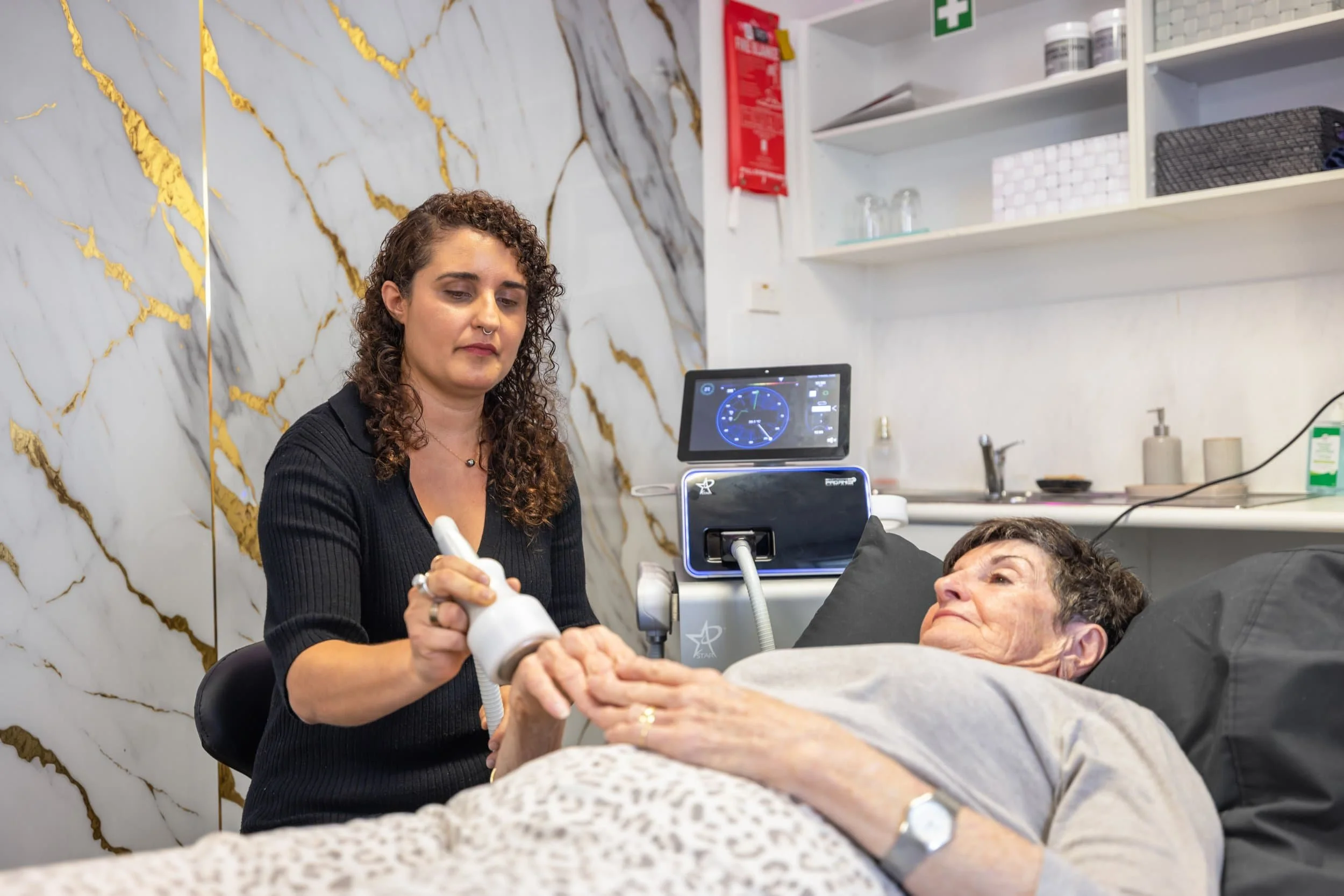 A healthcare professional administering ultrasound therapy to an elderly woman lying in bed in a medical facility. The woman is holding the caregiver's hand and looking at her. Medical equipment and supplies are visible in the background.