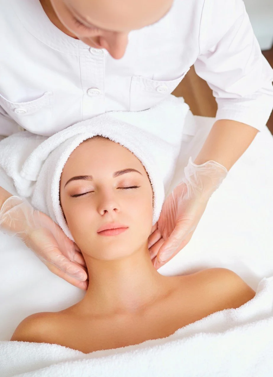 A woman receives a facial massage at a spa, with a towel wrapped around her head and a relaxed expression.