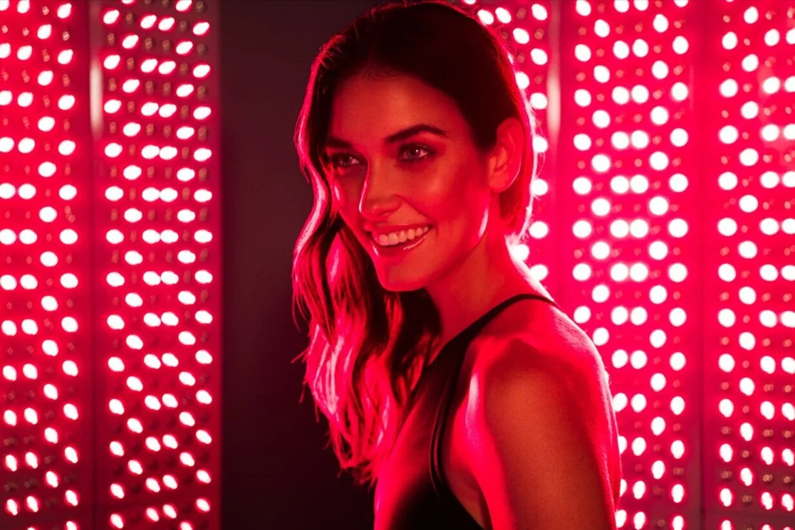 A woman with long wavy hair smiling, illuminated by a red light, standing in front of a background with red LED lights.