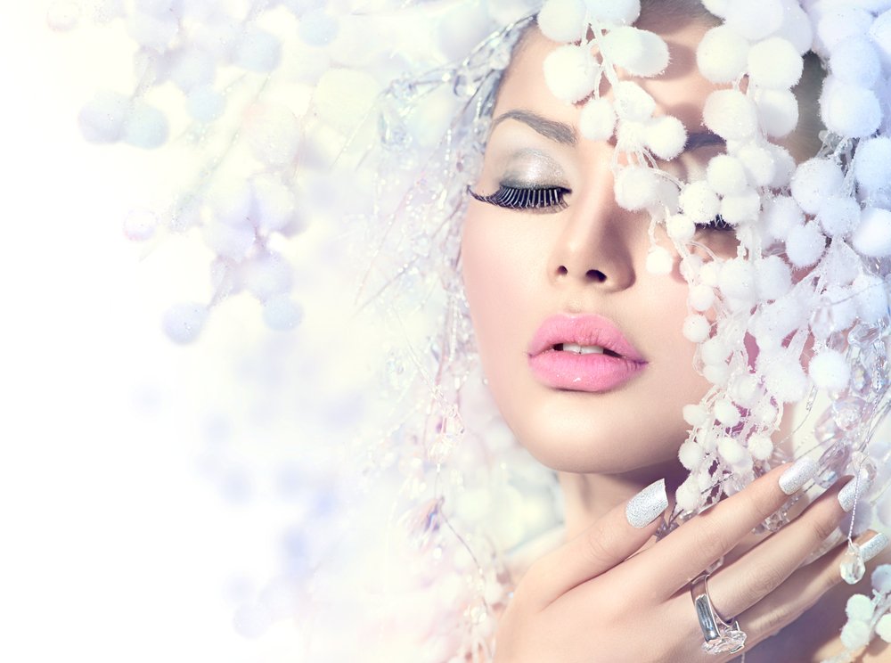 A woman with pink lipstick and silver eye makeup poses behind white, fluffy branches, with her eyes closed and hand near her face.