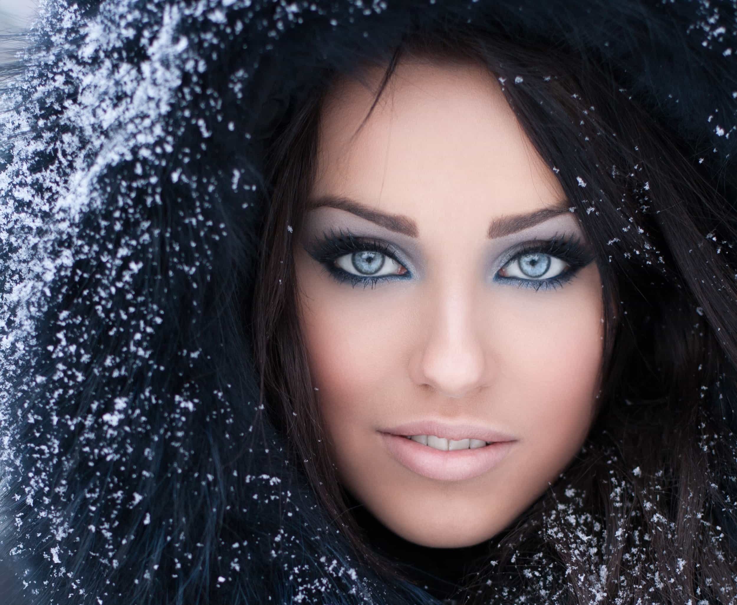 Close-up of a woman with icy blue eyes and dark hair, wearing a black fur-lined hood with snowflakes on it.