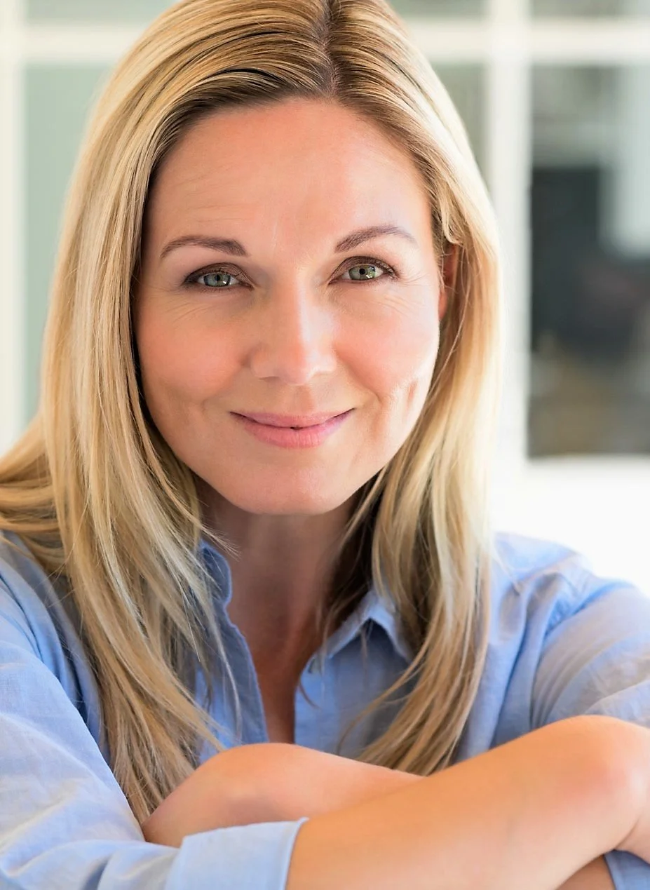 Close-up portrait of a smiling woman with long blonde hair wearing a light blue shirt, sitting indoors with a blurred background.