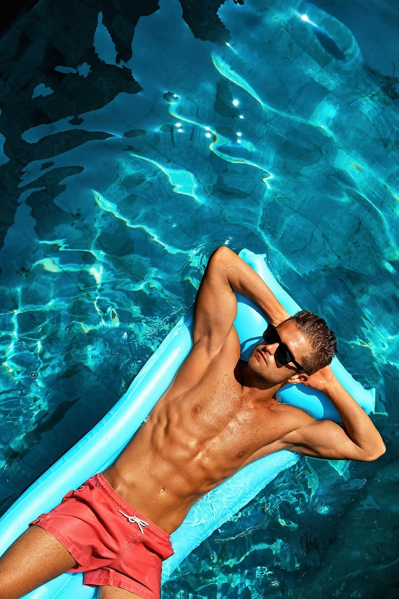 A man with a muscular build and sunglasses relaxing on a blue inflatable mattress in a swimming pool, wearing red swim trunks.