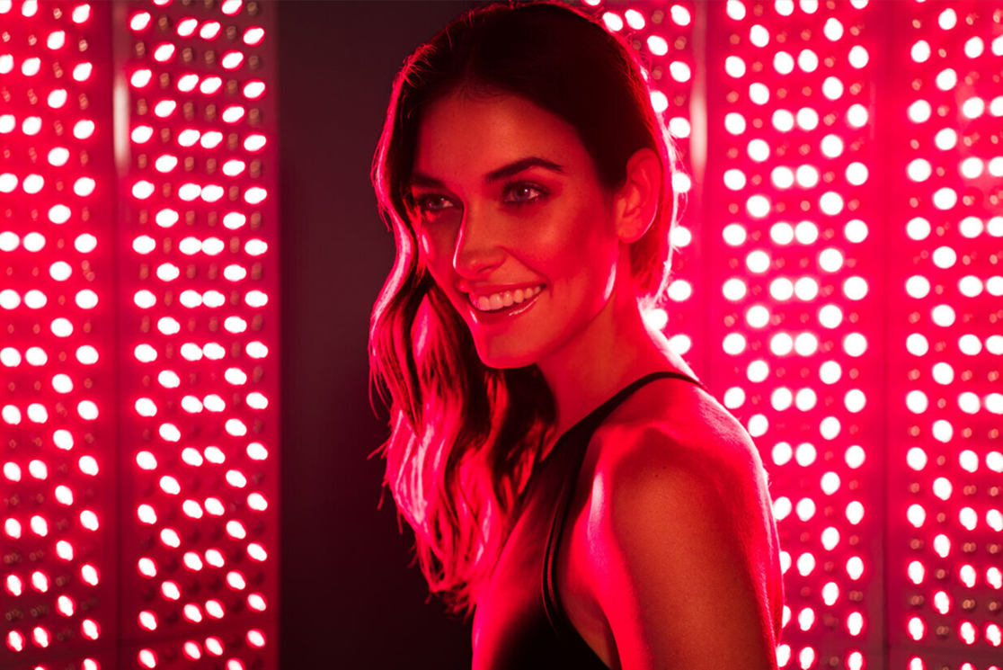 A woman with long wavy hair smiling, in front of red LED lights, wearing a black sleeveless top.
