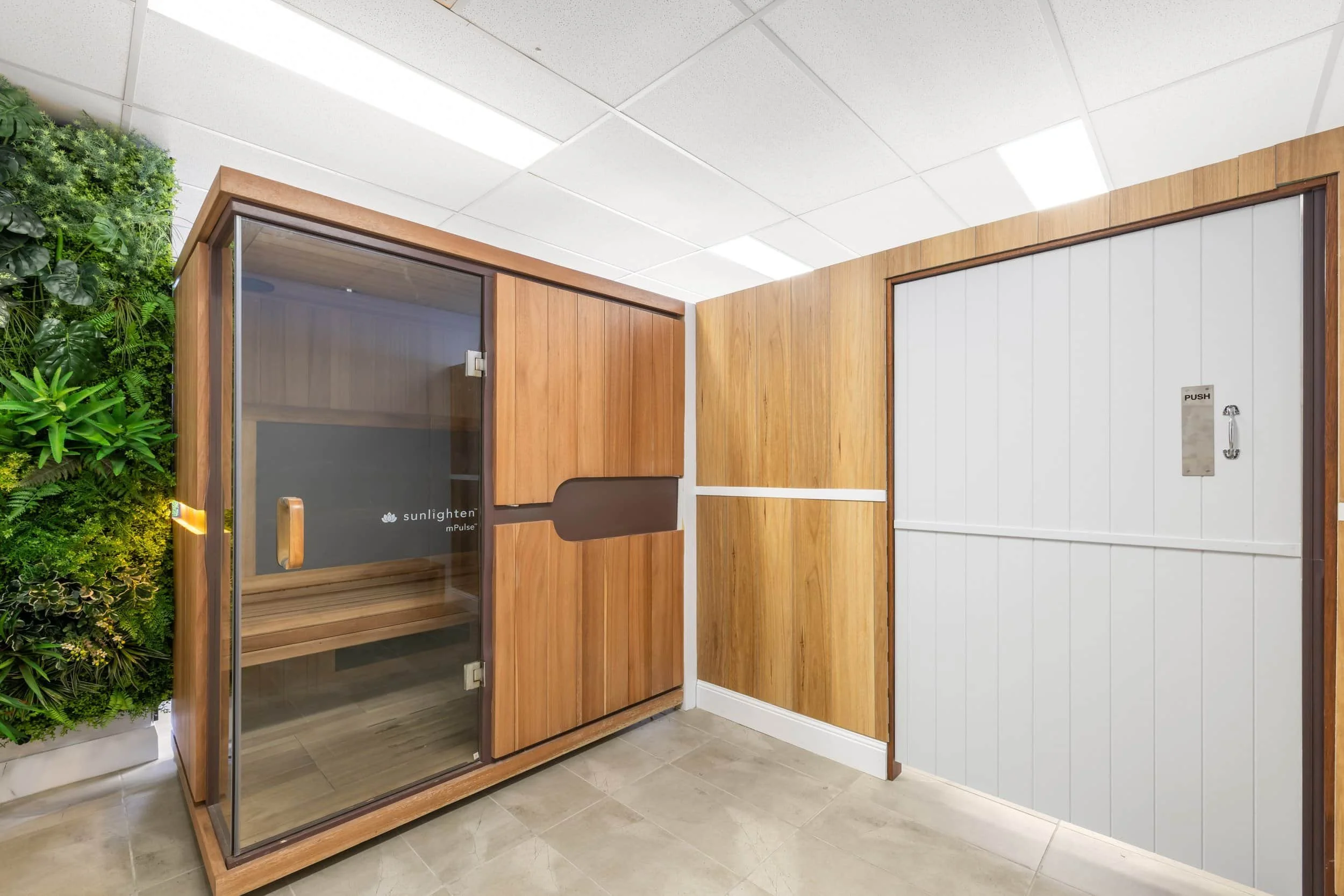 Indoor room with a sauna, featuring a wooden exterior, glass door, and adjacent wooden wall panels. Part of a green plant wall is visible on the left, and the ceiling has white tiles with fluorescent lighting.