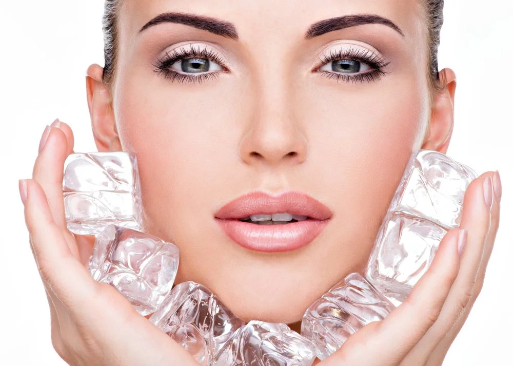 A woman with fair skin and makeup holding ice cubes near her face.