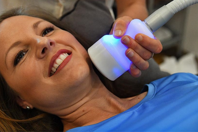 A woman lying down, smiling, receiving a facial treatment with a device emitting blue light.