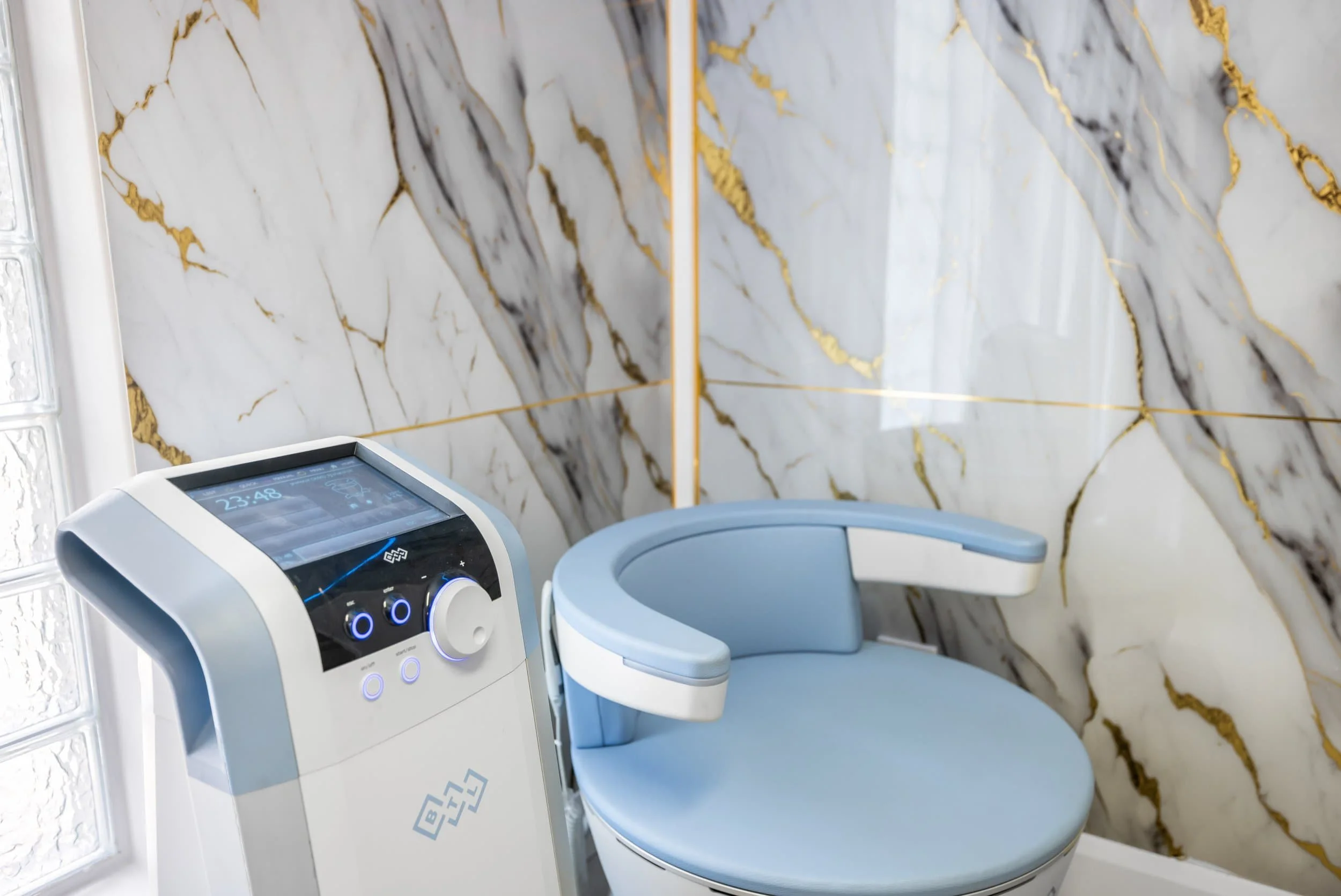 Emsella chair with control panel for incontinence care in a room with white marble walls and gold veins.