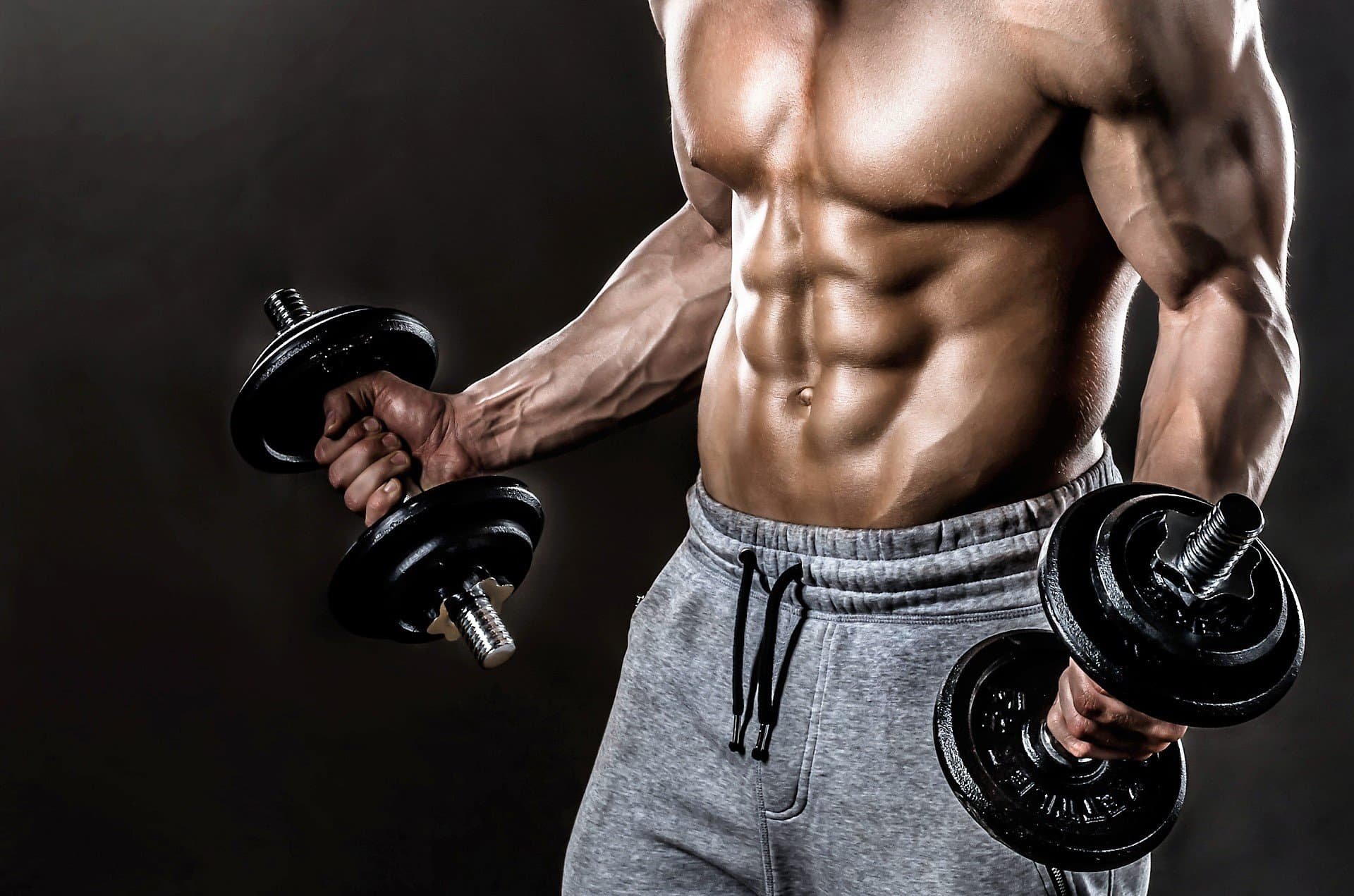 A muscular man with defined abs and arms, wearing gray sweatpants, holding dumbbells in both hands, against a dark background.