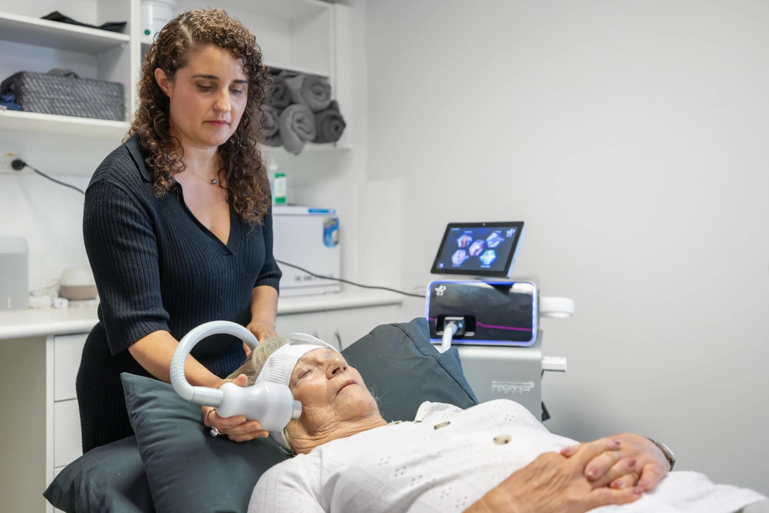 A woman lying on a medical bed receiving a treatment with a handheld device, while another woman stands beside her, operating the device. The setting appears to be a clinical or medical environment with medical equipment and a computer in the backgro