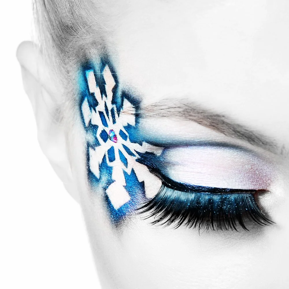 Close-up of a woman's eye with artistic snowflake makeup around the temple, featuring blue, white, and purple colors, and dramatic eyelashes.