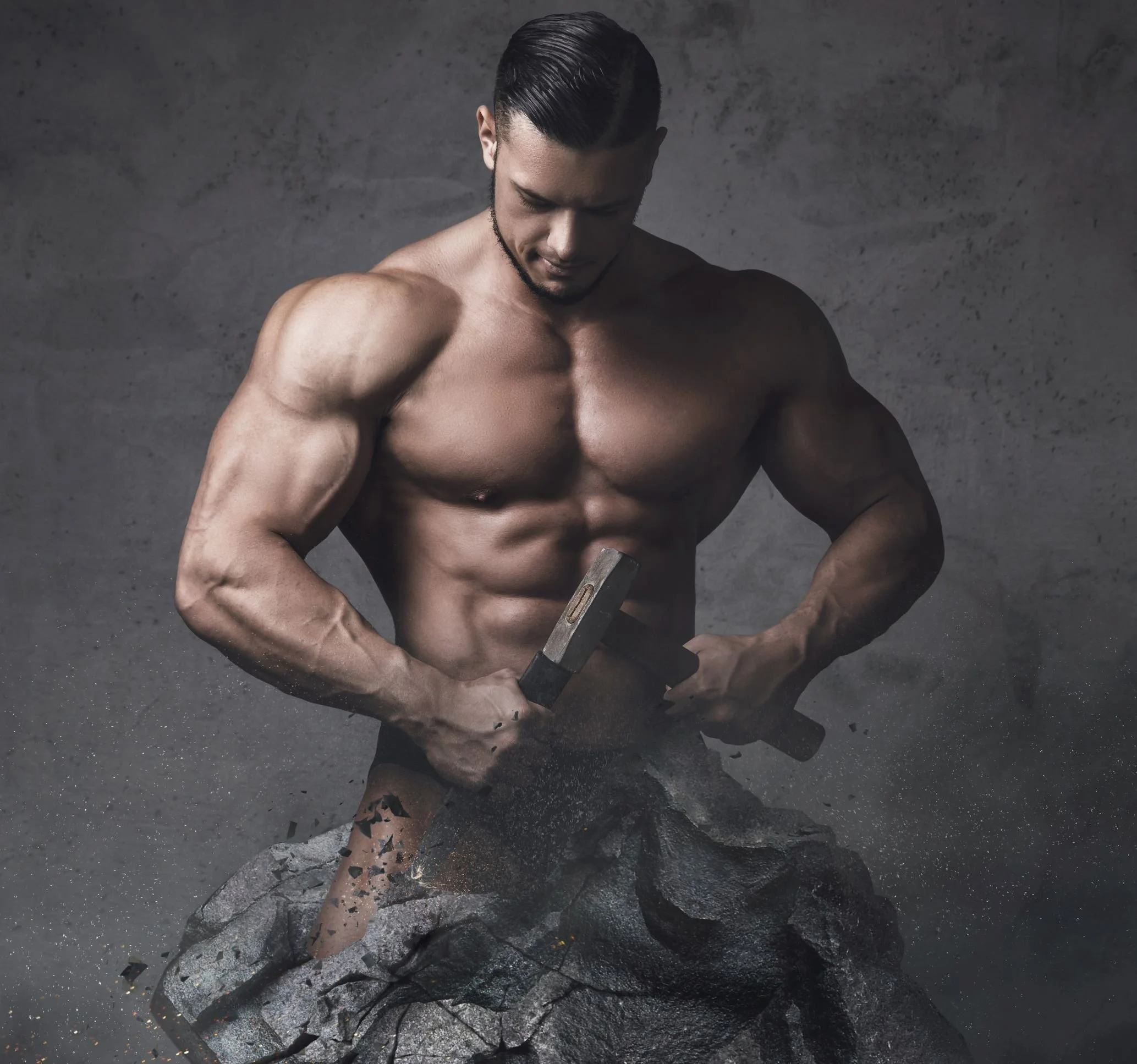 A muscular man with slicked black hair standing shirtless behind a cracked rock, holding a hammer in both hands, with a gray textured background.