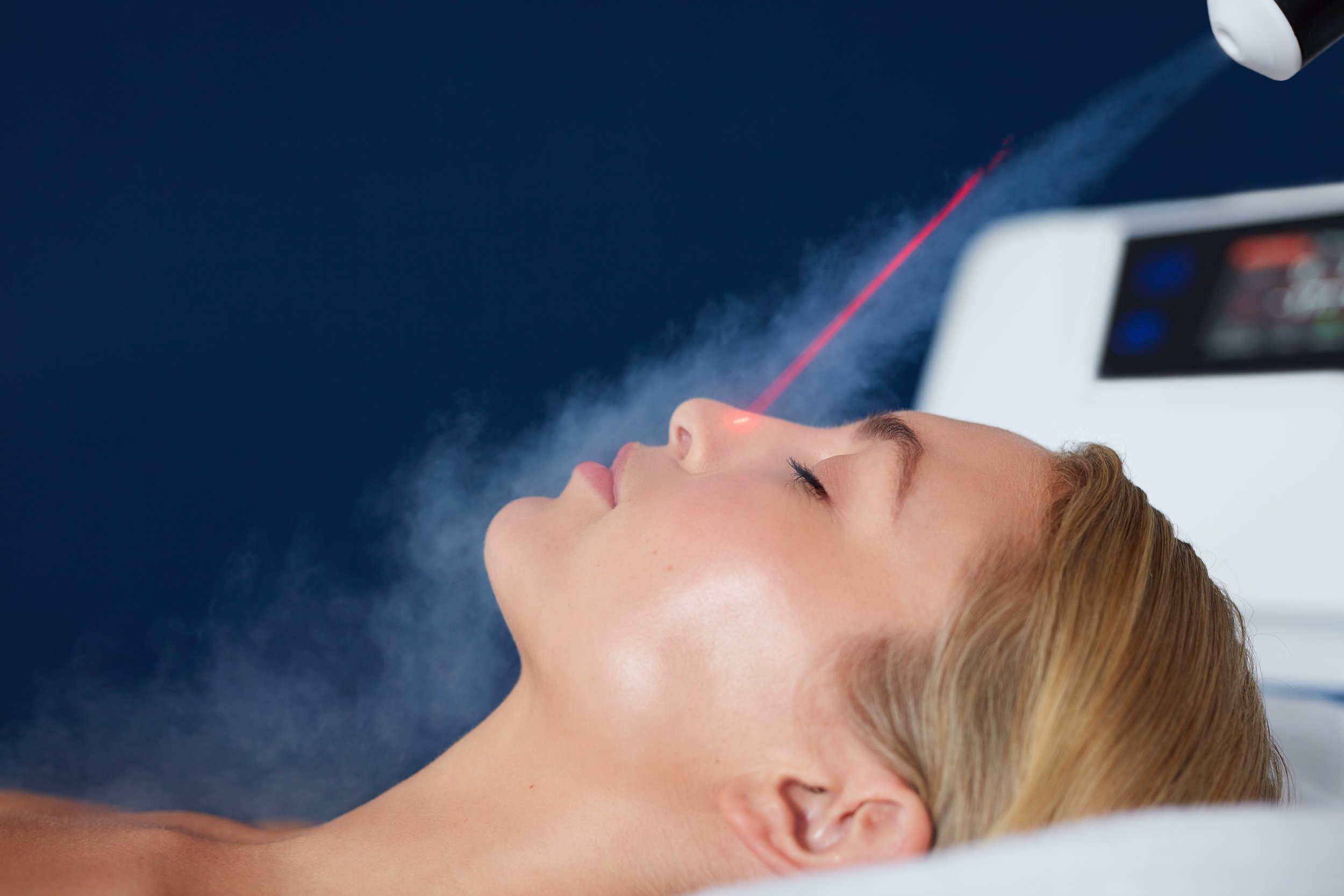 A woman lying eyes closed during a laser treatment, with a device emitting a red laser beam on her face.