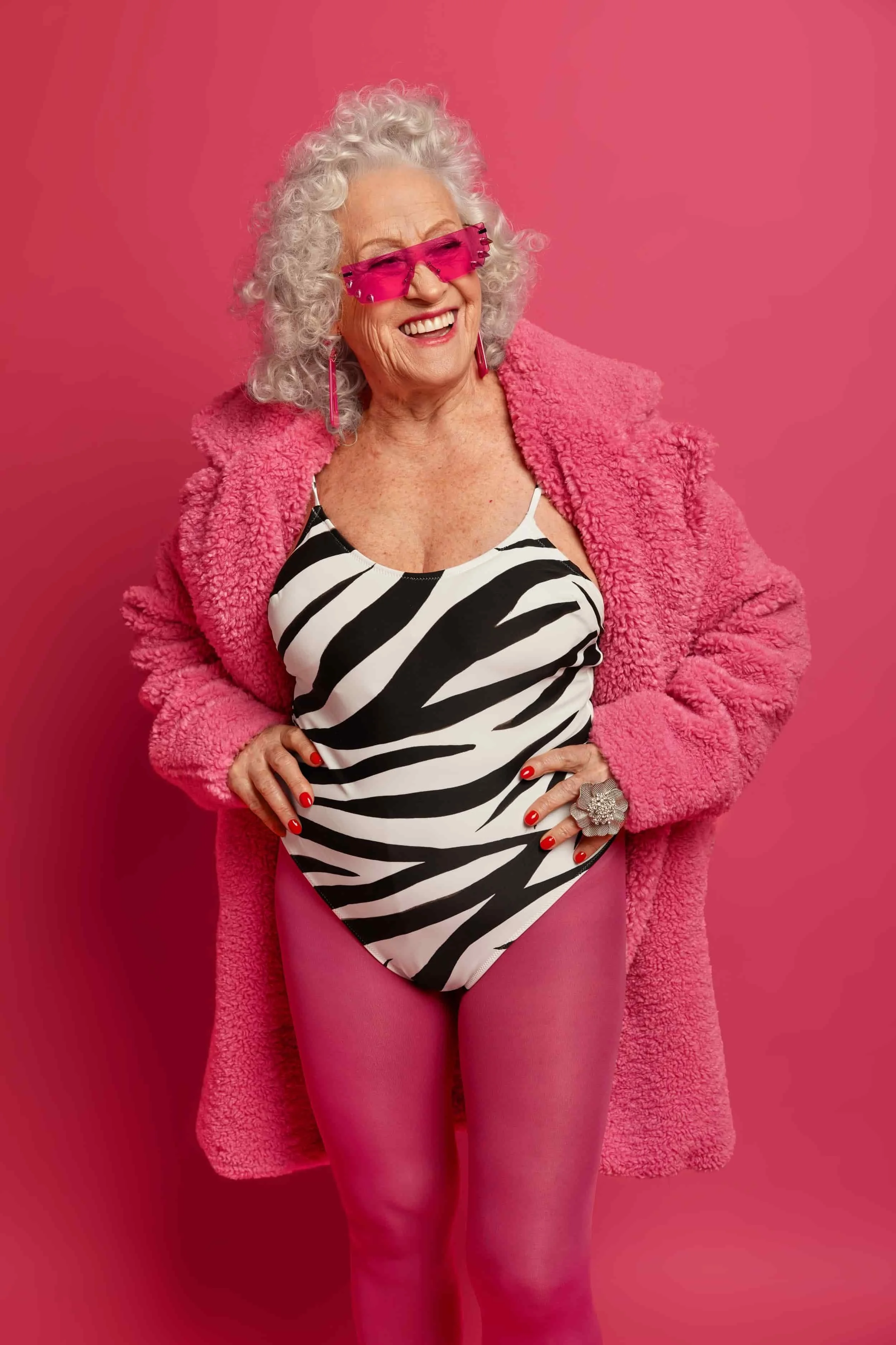 An elderly woman smiling, wearing pink sunglasses, a pink fuzzy coat, a black and white zebra print swimsuit, pink tights, and statement jewelry, standing against a pink background.