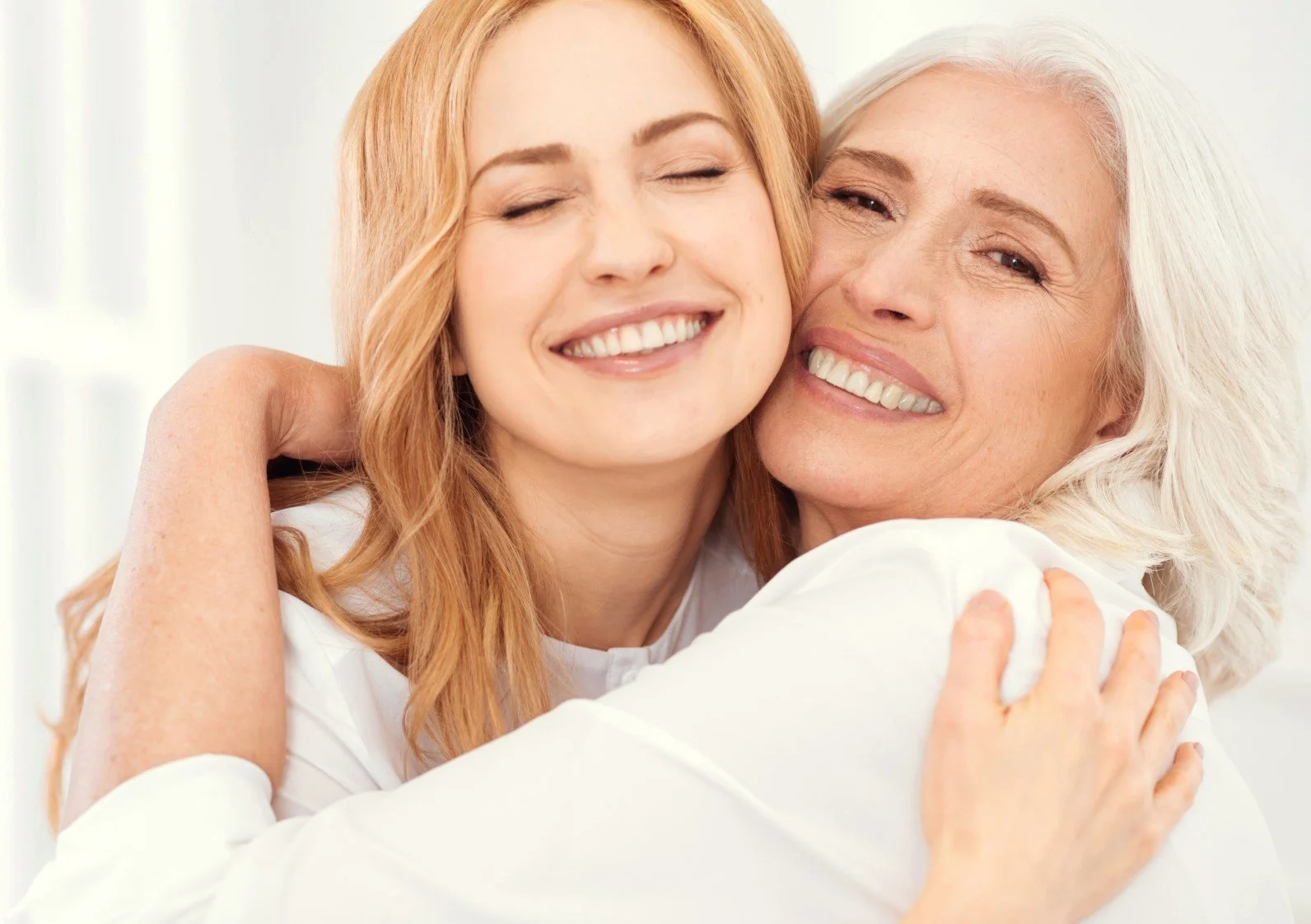 A young woman and an older woman hugging and smiling with their eyes closed in a bright, happy setting.