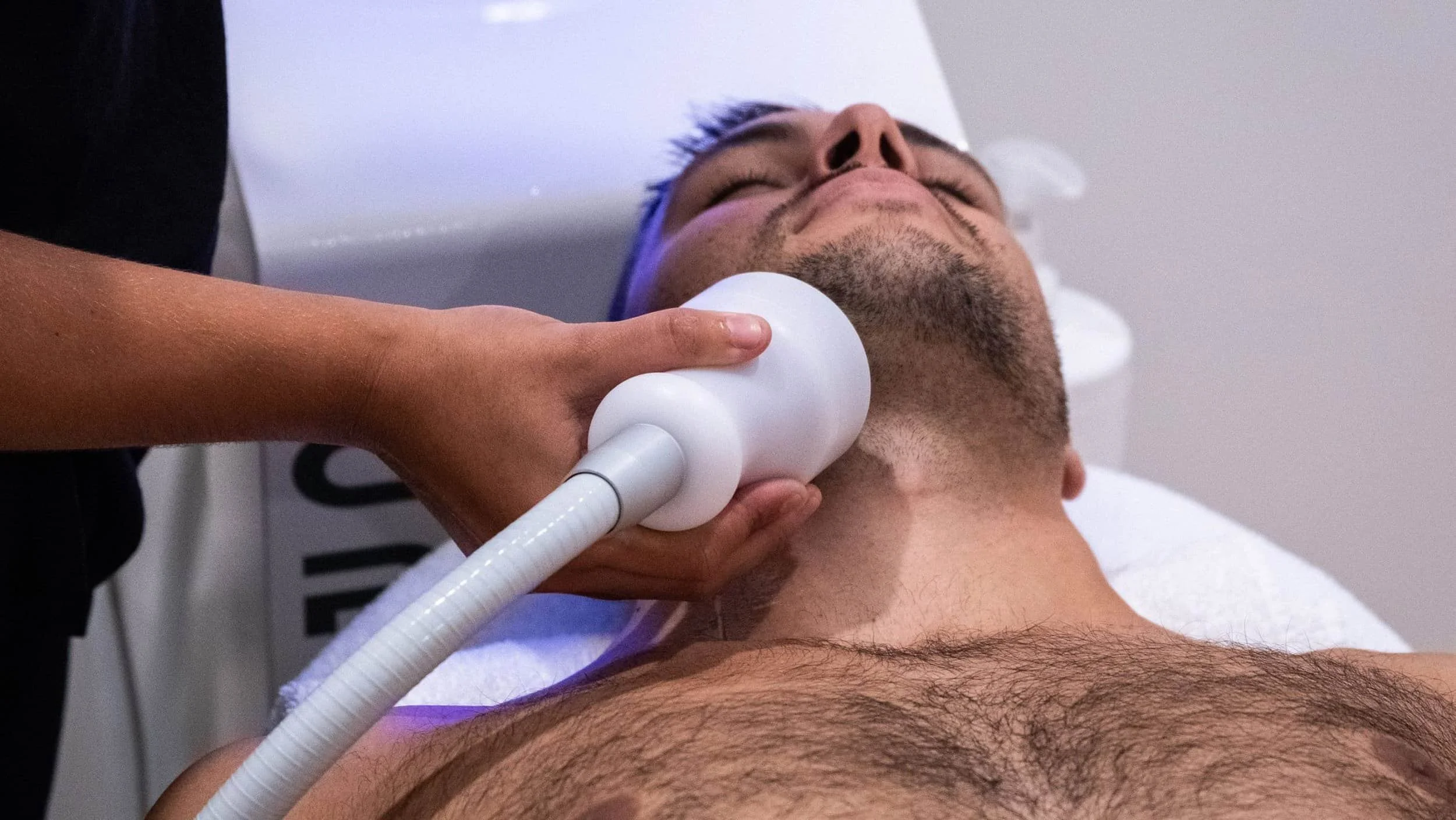 A man receiving a facial treatment with a handheld device on his neck while lying down, eyes closed, in a clinical setting.