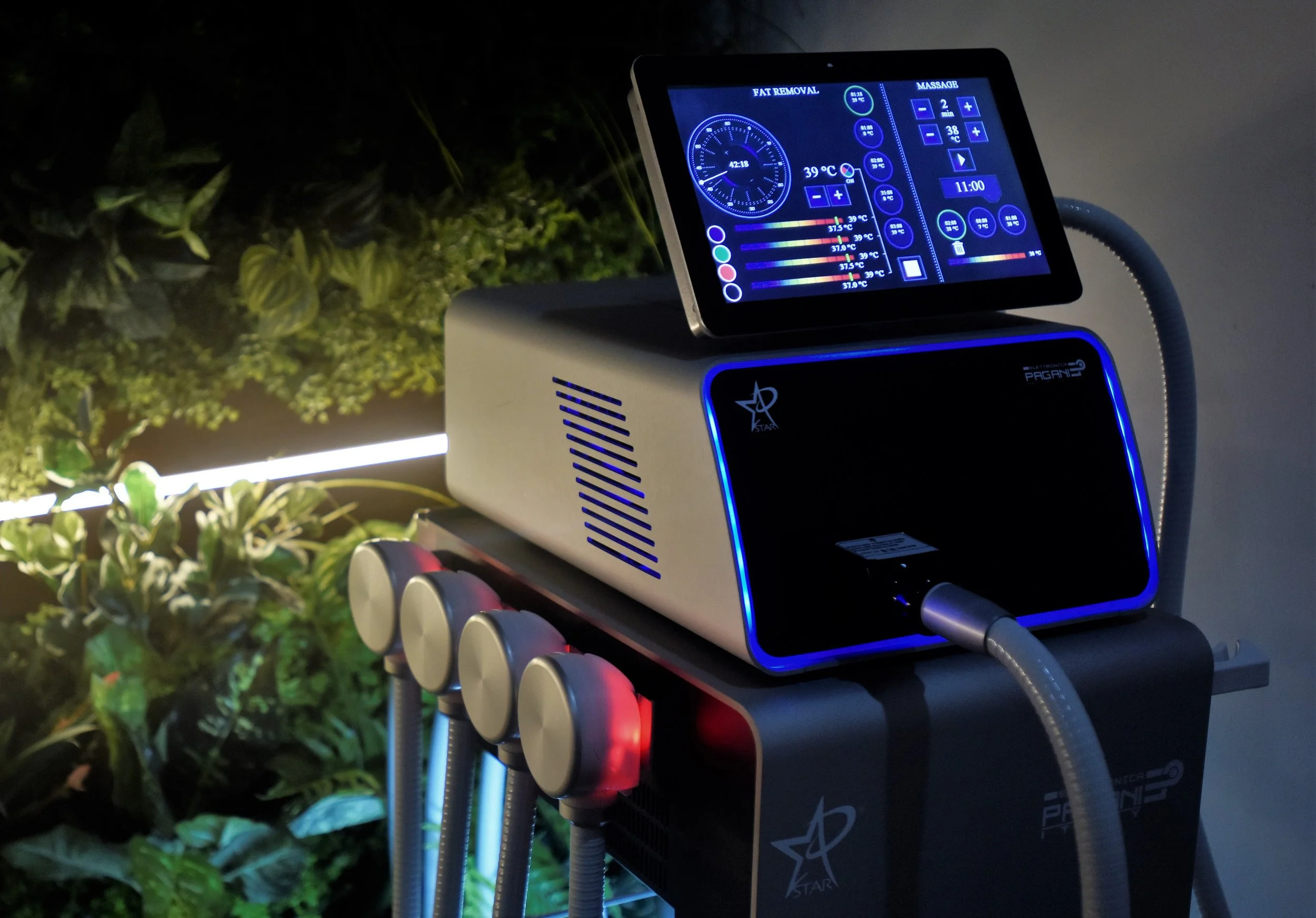 A medical cosmetic device with a digital screen showing temperature settings, timers, and controls, adjacent to a row of four round applicators, connected by flexible tubes, placed in front of a wall with green plants.