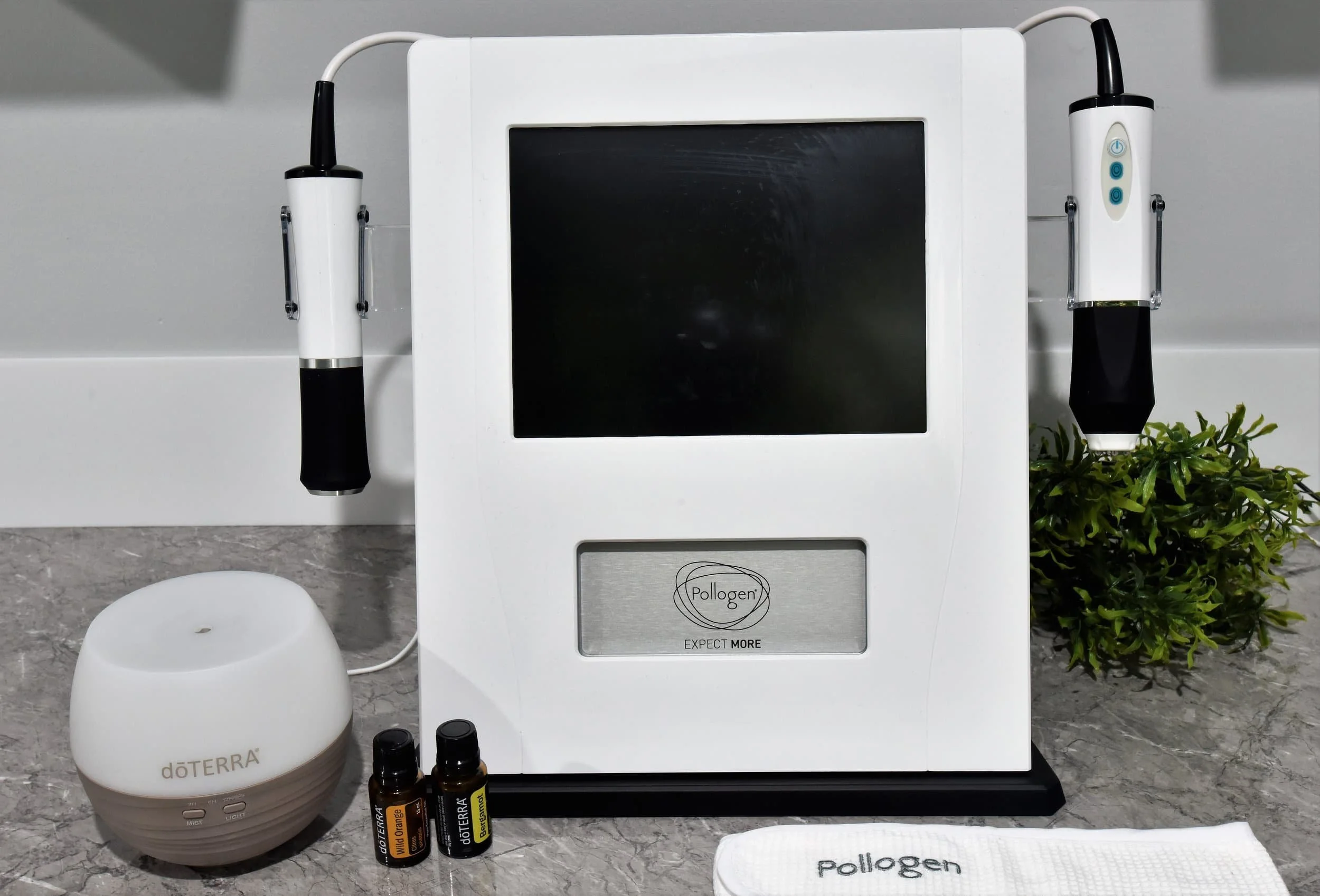 A Pollogen microneedling device with two handpieces attached, a white diffuser with the doTERRA logo, two essential oil bottles, a Pollogen towel, and a potted plant on a marble surface.