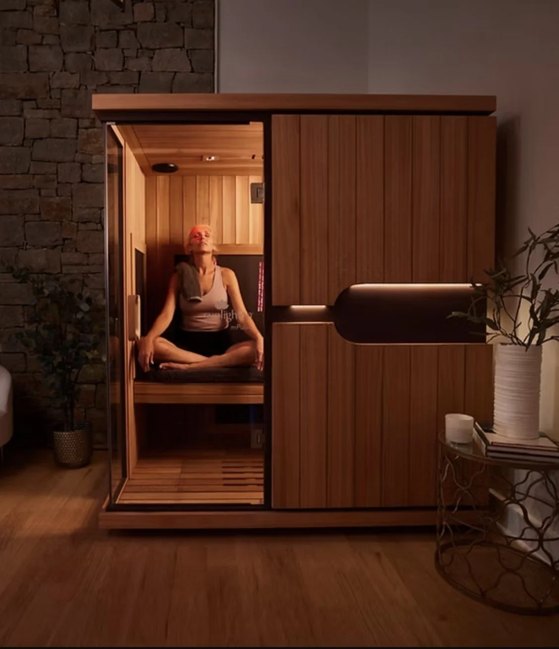 A woman practicing yoga in a wooden infrared sauna at home, sitting cross-legged with eyes closed.