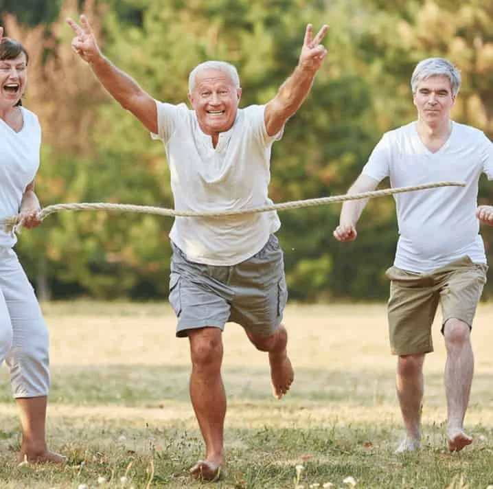 Older man winning a three-legged race with two younger men outdoors in a park, all smiling and holding a rope.