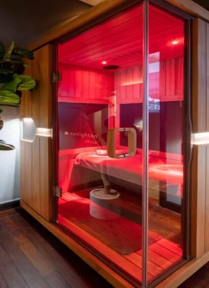 A red-colored infrared sauna with glass panel door, located indoors in a room with wood flooring and walls, and a potted plant nearby.
