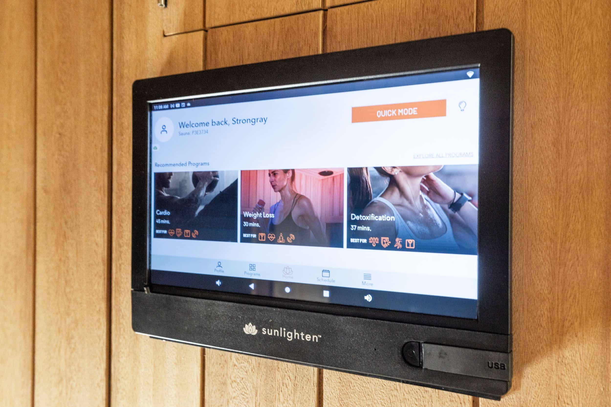 A touchscreen control panel mounted on a wooden wall, displaying various fitness programs such as cardio, weight loss, and detoxification at Strongray wellness center.