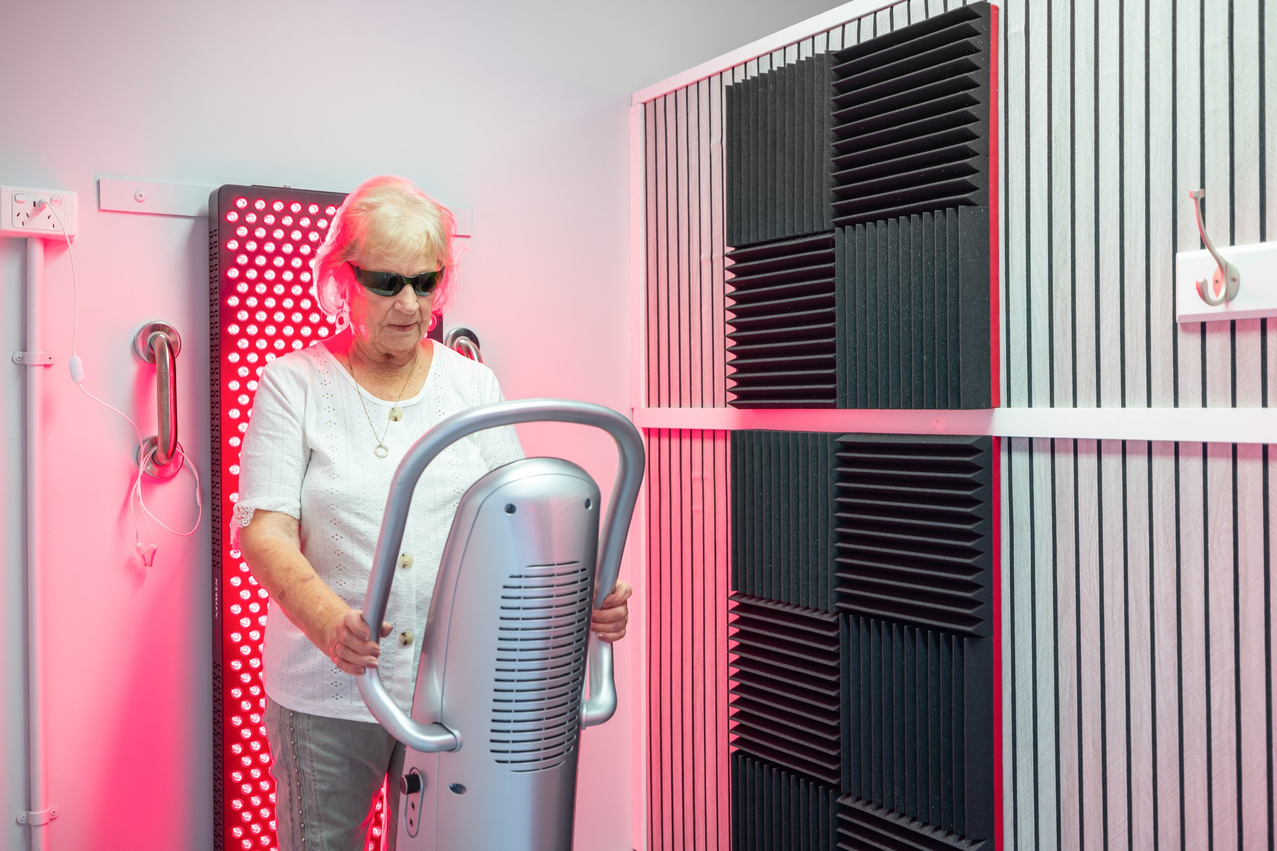 Older woman in sunglasses and white top using a light therapy device in a room with soundproof foam panels.