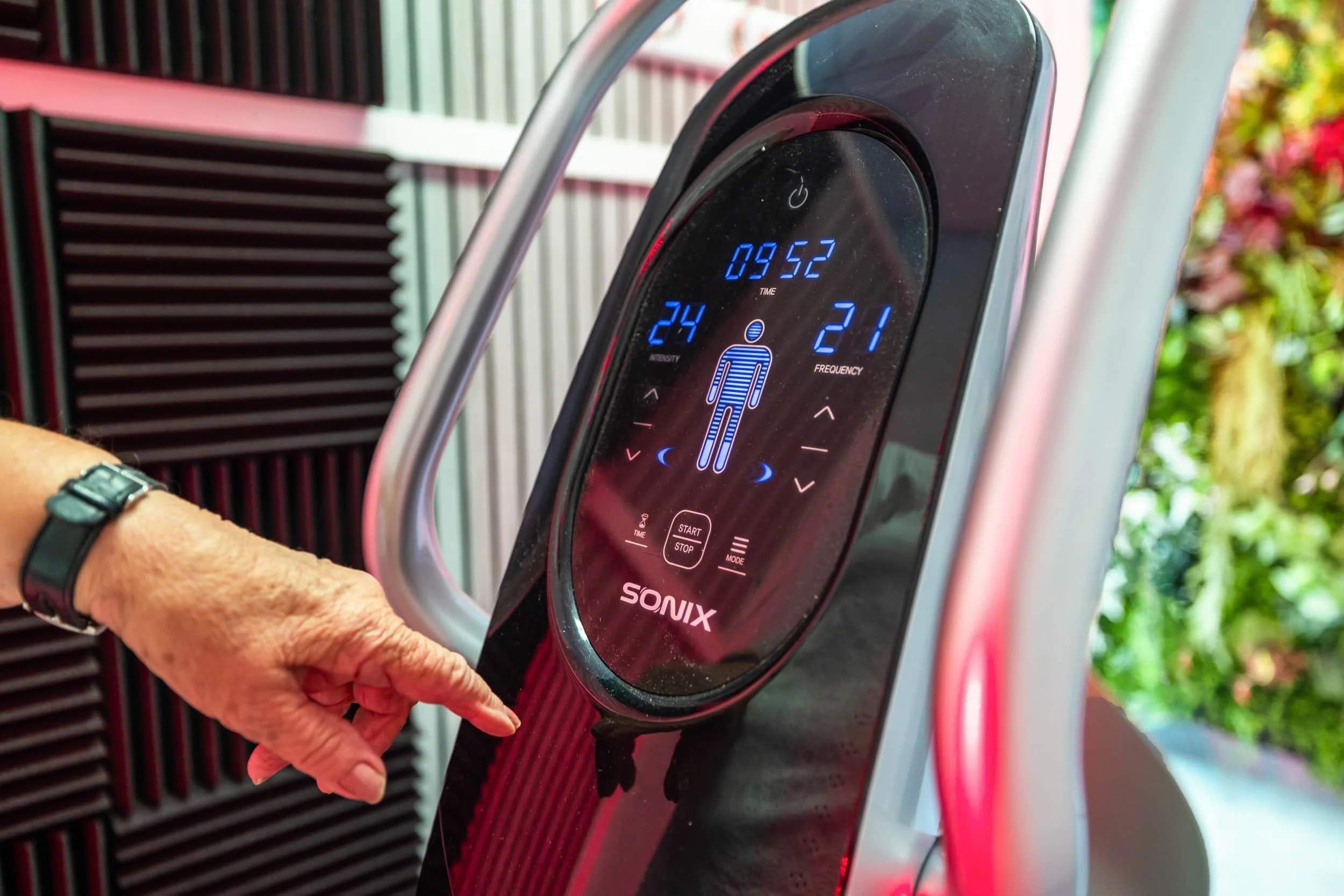 Digital health and wellness monitor of a SONIX Whole Body vibration & massage device with a display showing settings, a person figure, and , displaying metrics such as time, intensity, and frequency, and a person's hand pressing a button.