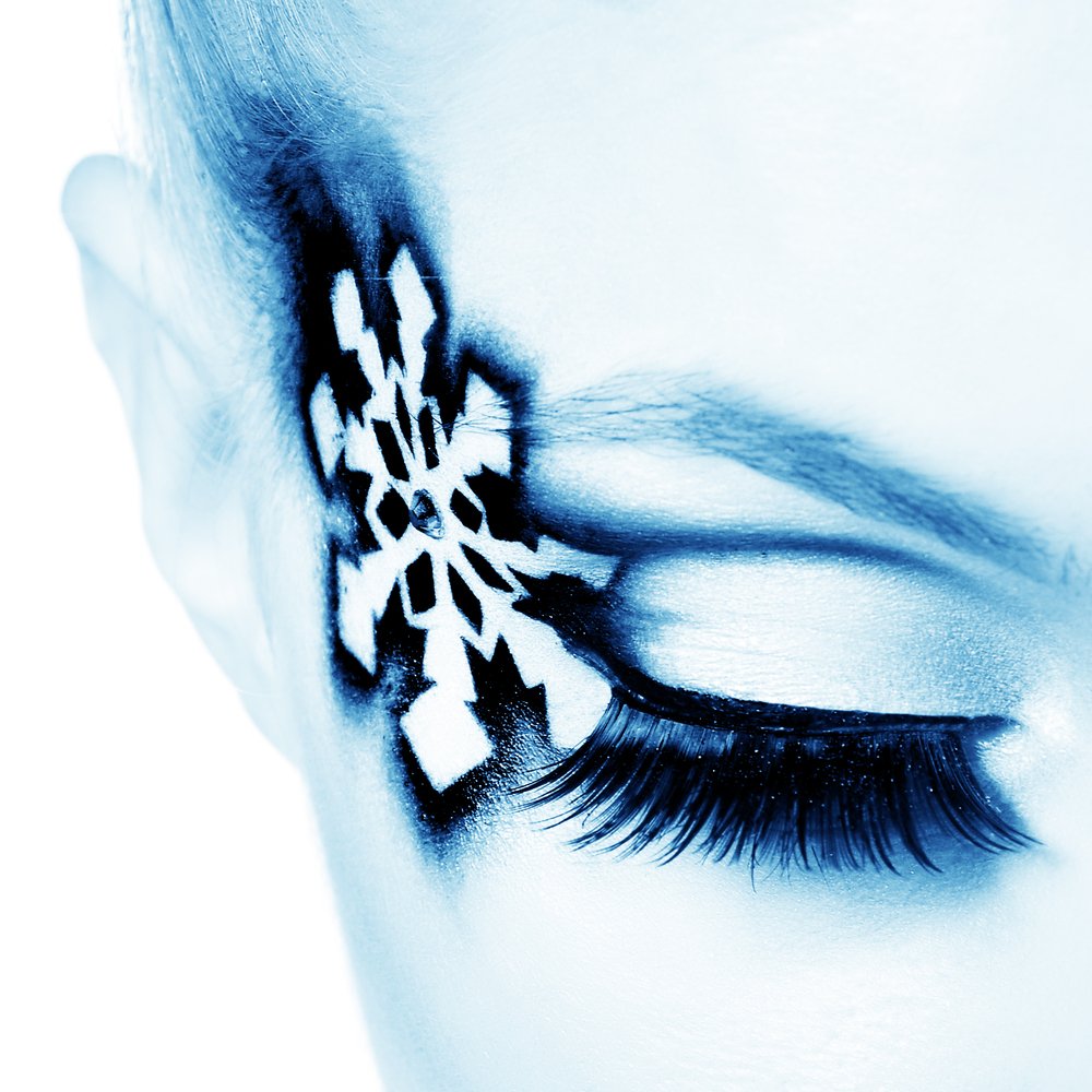 Close-up of a woman's face with artistic ice crystal snowflake design on cheek near eye, long eyelashes, and eye makeup with cool blue tones.