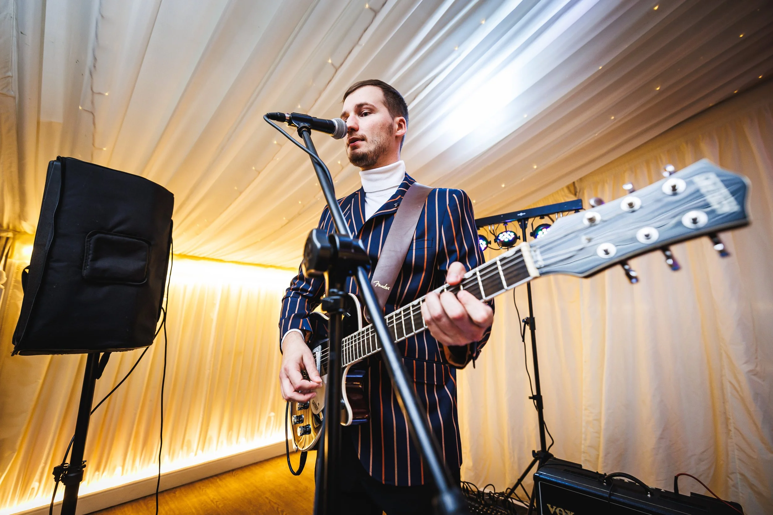A man in a striped blazer playing an electric guitar and singing into a microphone at a music event or party, with stage lighting and sound equipment in the background.