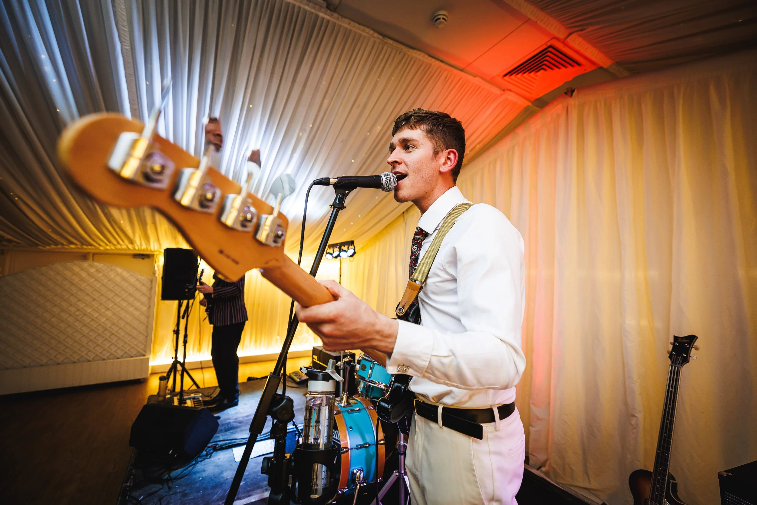 A young man in a white shirt and tie playing an electric guitar and singing into a microphone, with a drummer and a bassist in the background at a live music event.