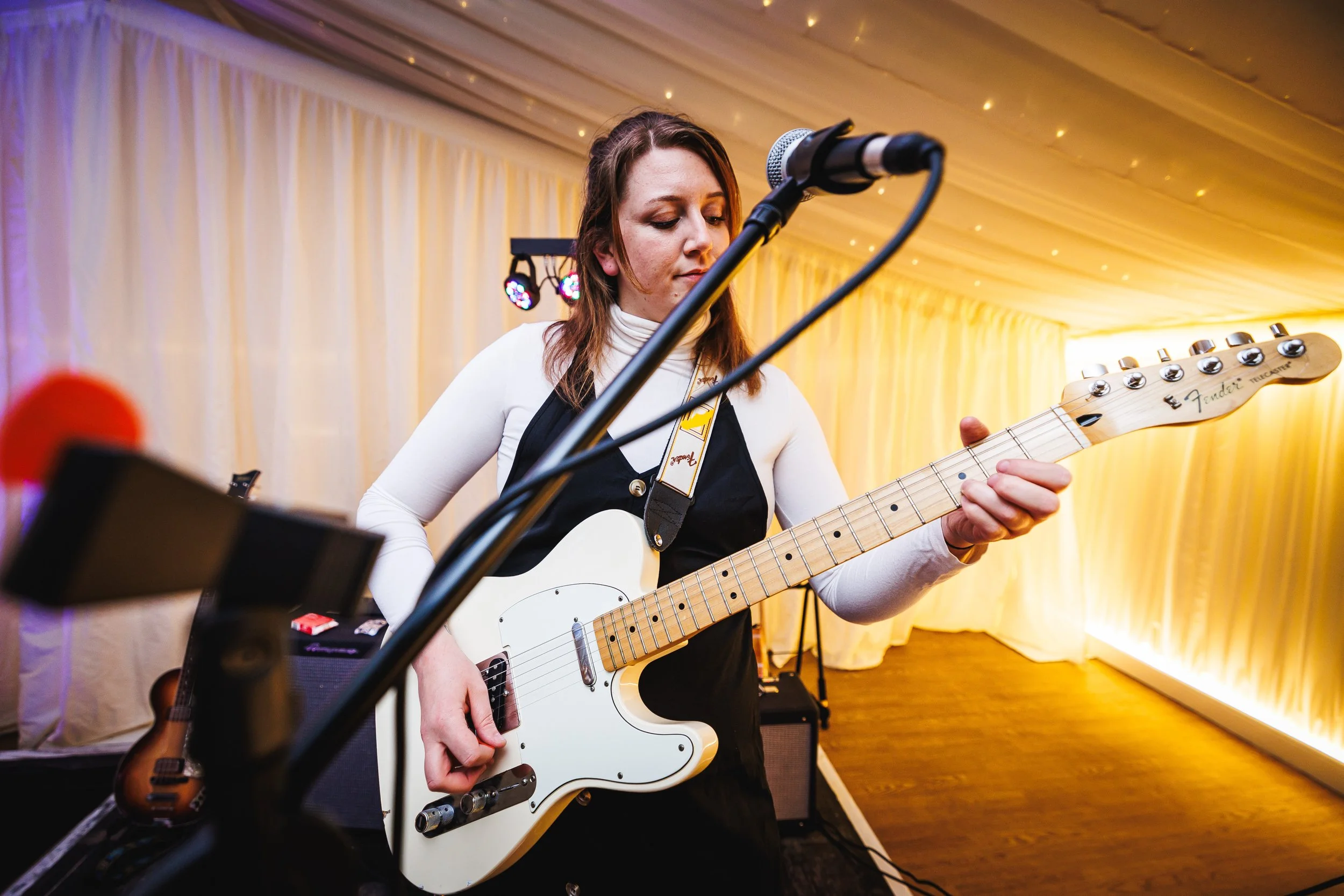 A woman playing an electric guitar at an indoor event, with a microphone in front of her and musical equipment in the background, illuminated with warm lighting.