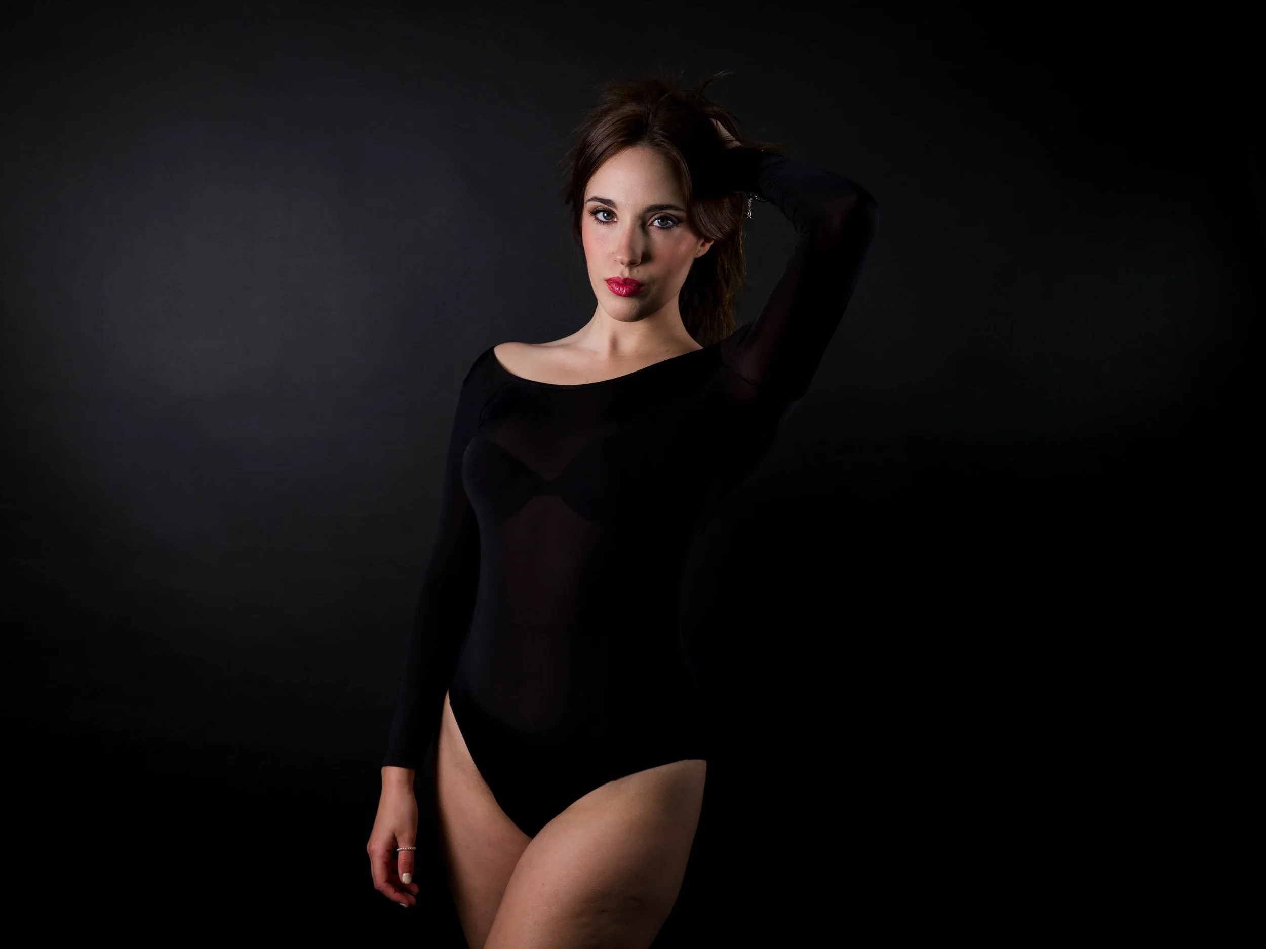 Mujer con cabello marrón oscuro y maquillaje con labios rojos, vestida con un body negro transparente, posando con una mano en la cabeza, contra fondo negro
