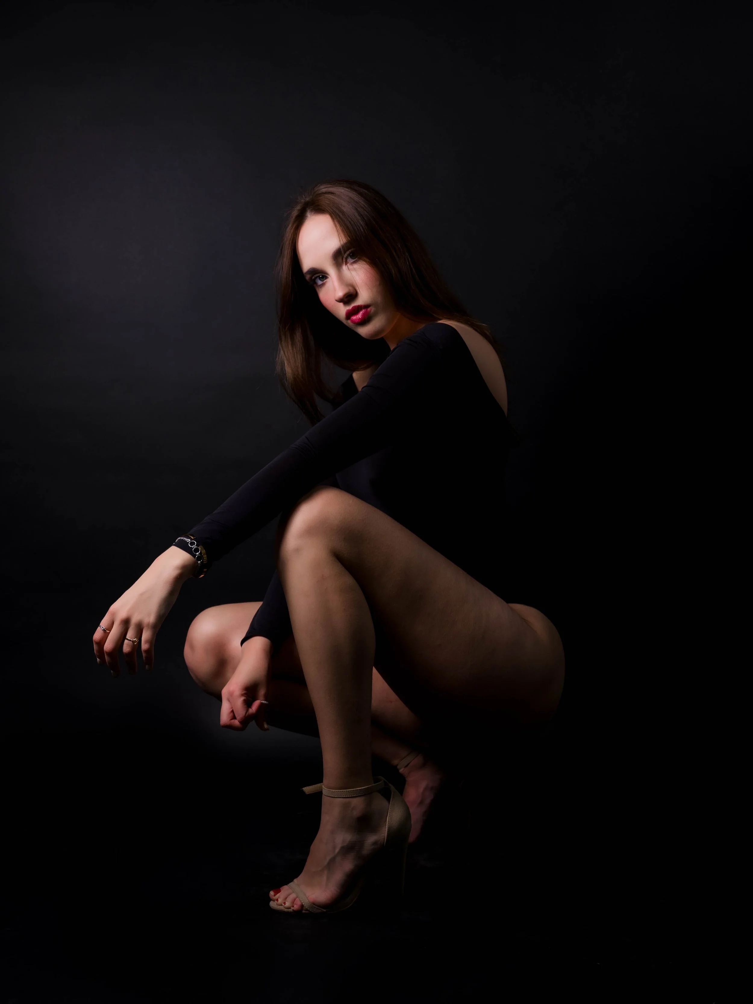Mujer joven con cabello castaño oscuro posando en un fondo negro, vestida con ropa negra, con un pie con tacón beige, en posición de cuclillas