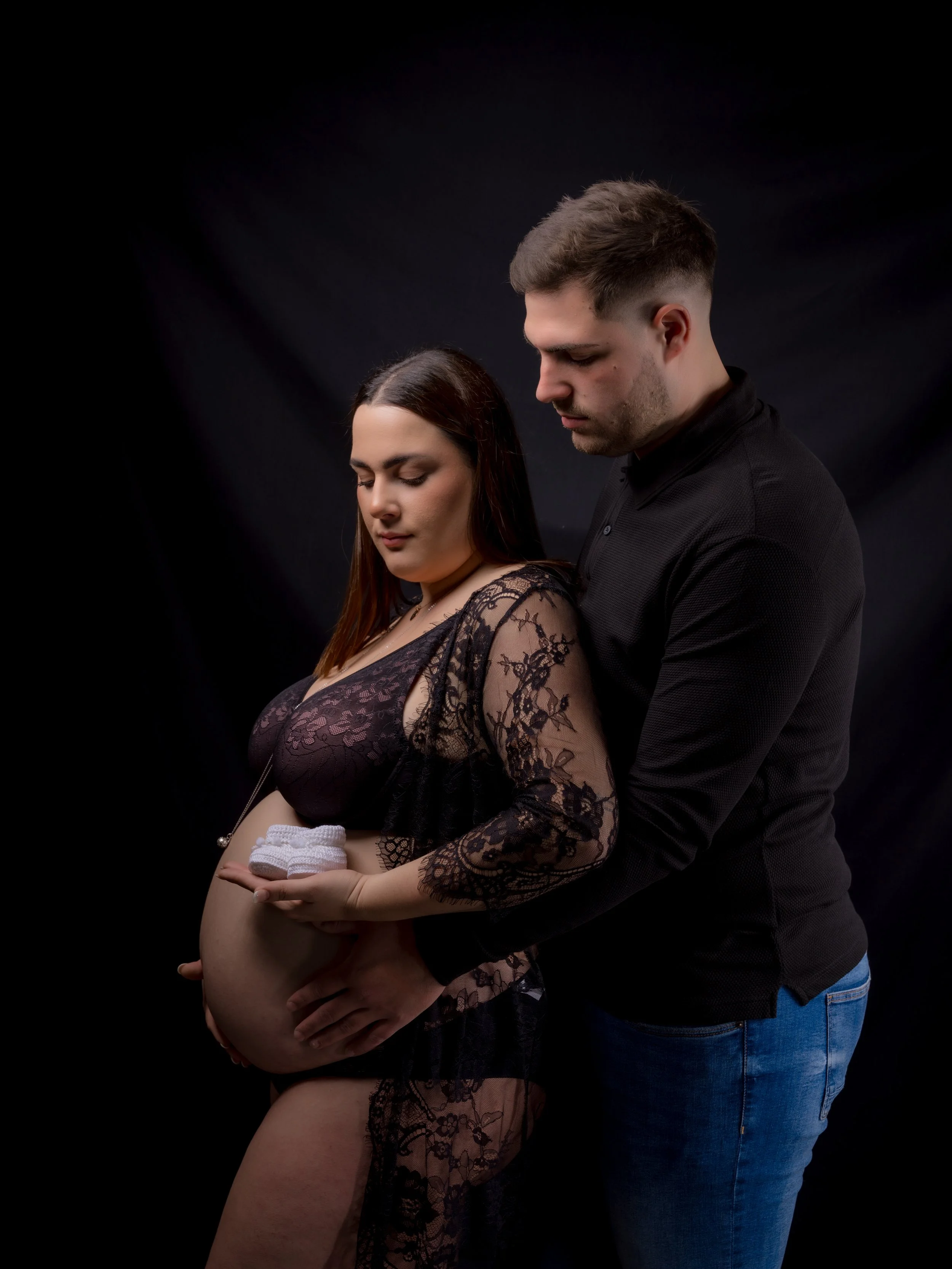 Una pareja mira la barriga de una mujer embarazada que sostiene en sus manos prendas blancas, ambos evitan explorar y muestran expresión de calma, en un fondo negro.