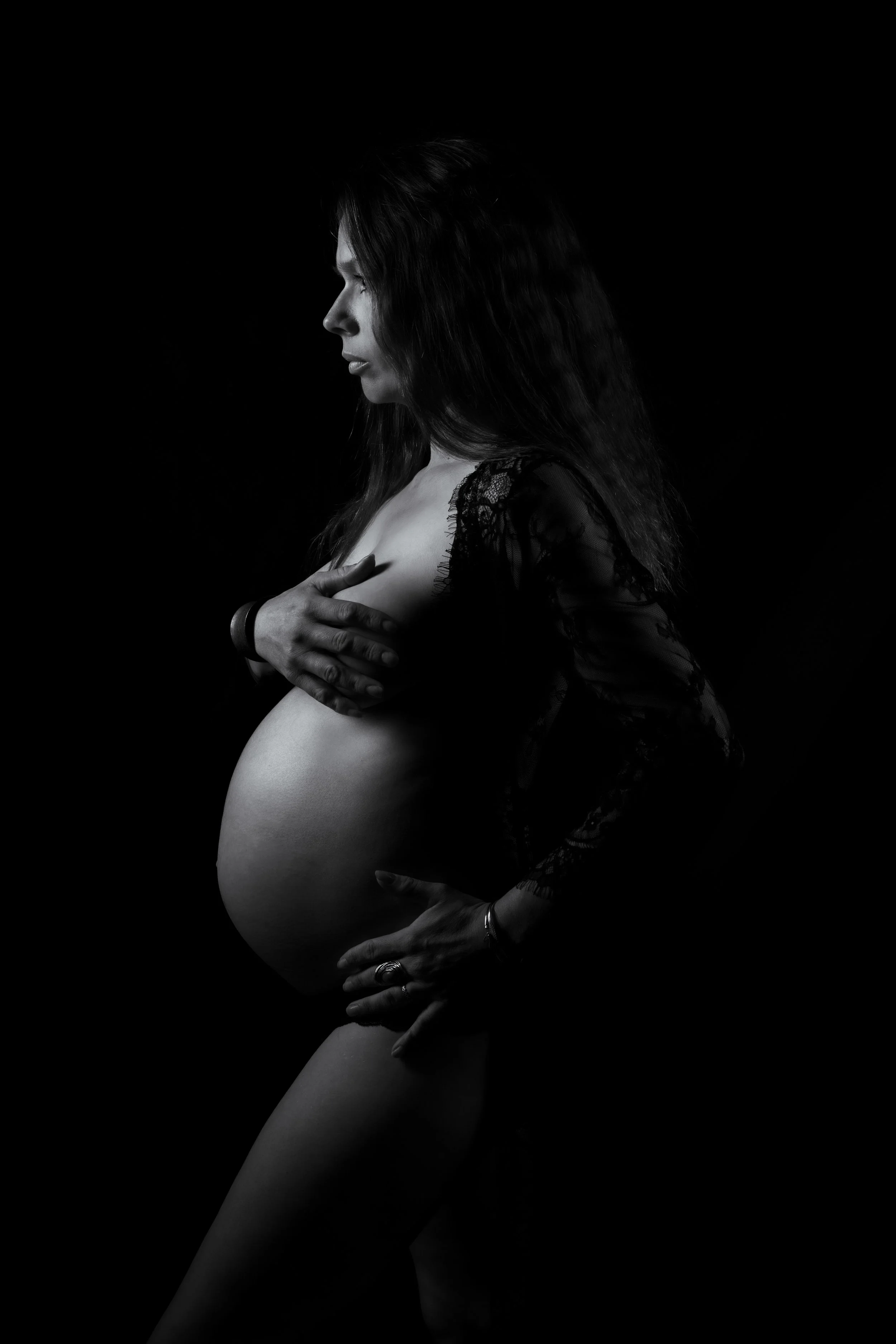 Sesion desnudo de embarazo en Sevilla, Bormujos y Aljarafe. Mujer embarazada en retrato en blanco y negro, de perfil, cubriéndose parcialmente con su mano, con cabello oscuro y vestido de encaje.