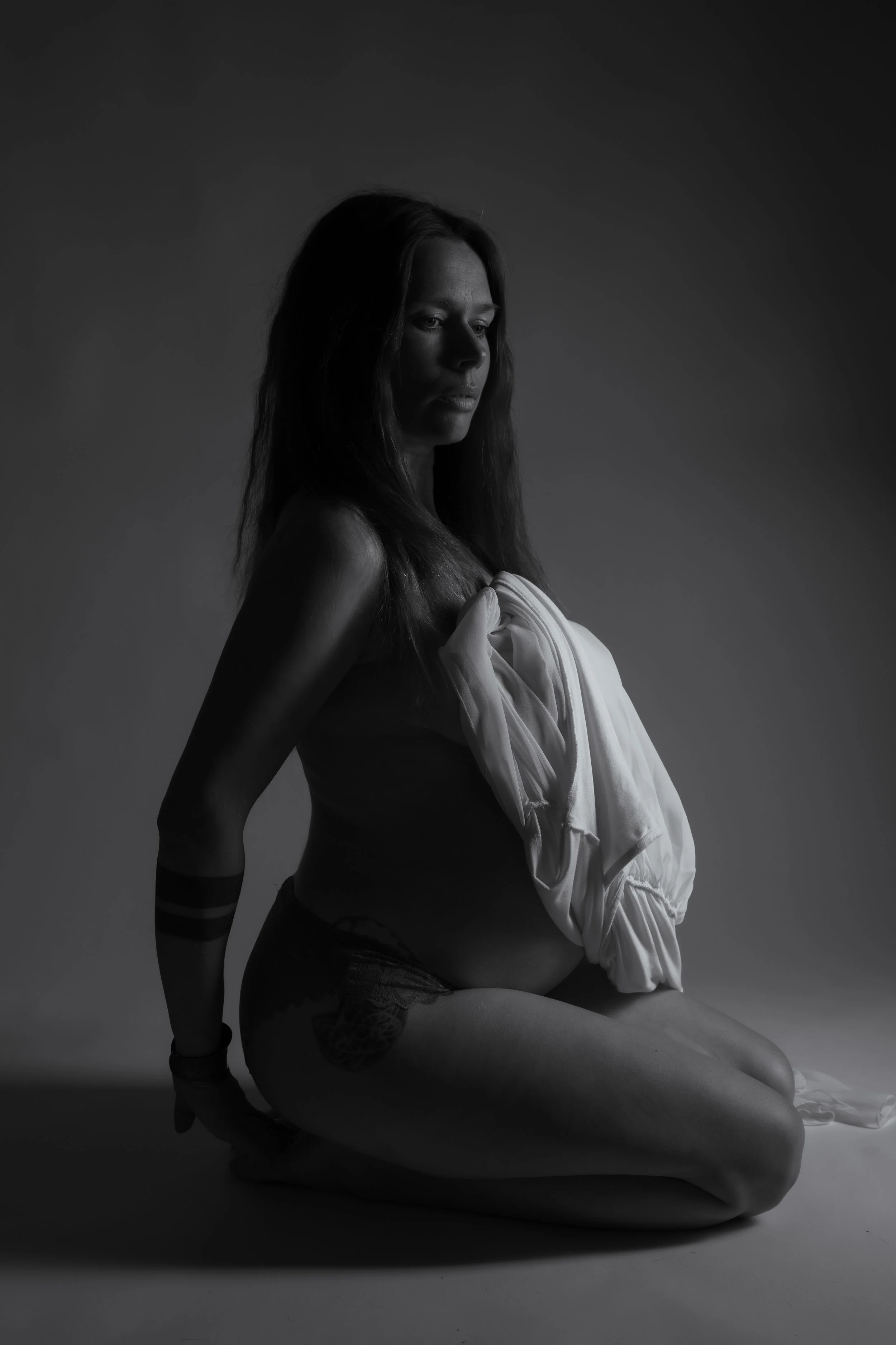 Estudio fotográfico en Sevilla, Bormujos. Mujer desnuda con ropa blanca enrollada sobre el pecho, sentada en posición de rodillas, en un fondo gris oscuro, en una fotografía en blanco y negro.