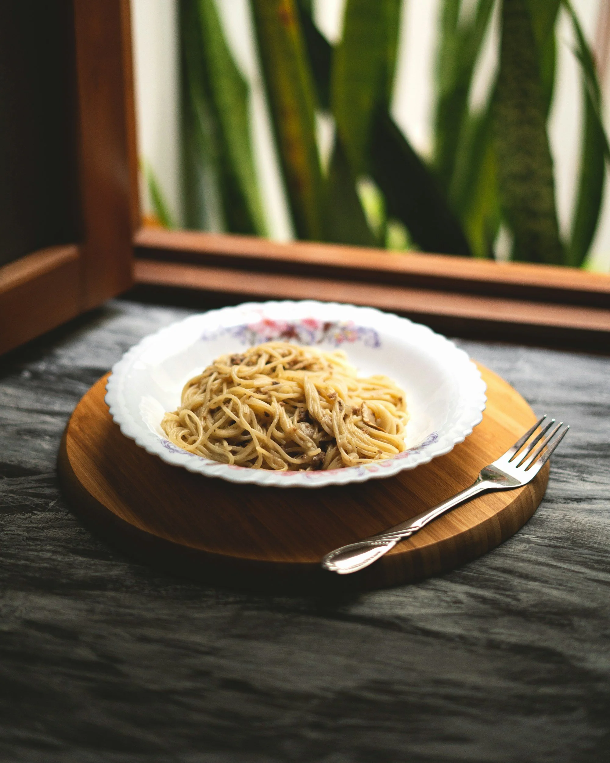Plato de espaguetis con salsa y carne, en un plato blanco con borde decorado, sobre una base de madera redonda, acompañado de un tenedor plateado, con fondo de ventana y plantas verdes.