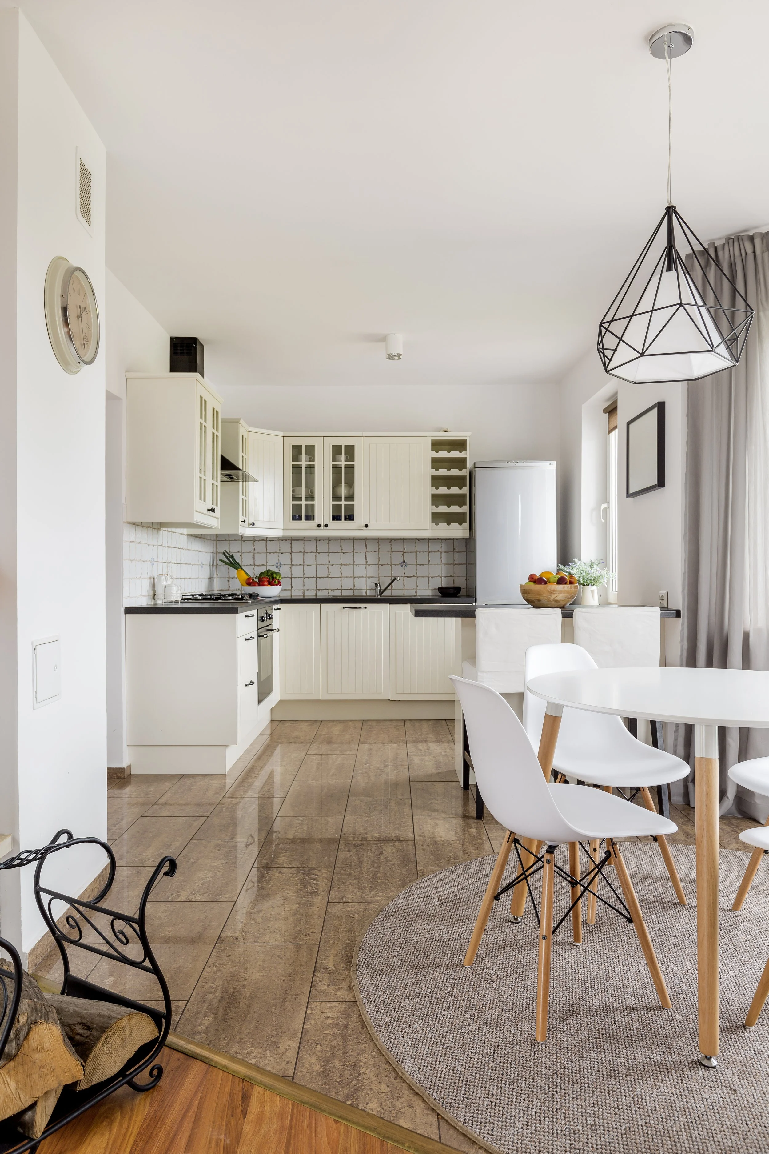 Cocina moderna en casa con armarios blancos, mesa redonda con sillas blancas de madera, área de comedor con lámpara decorativa y ventanas con cortinas grises.