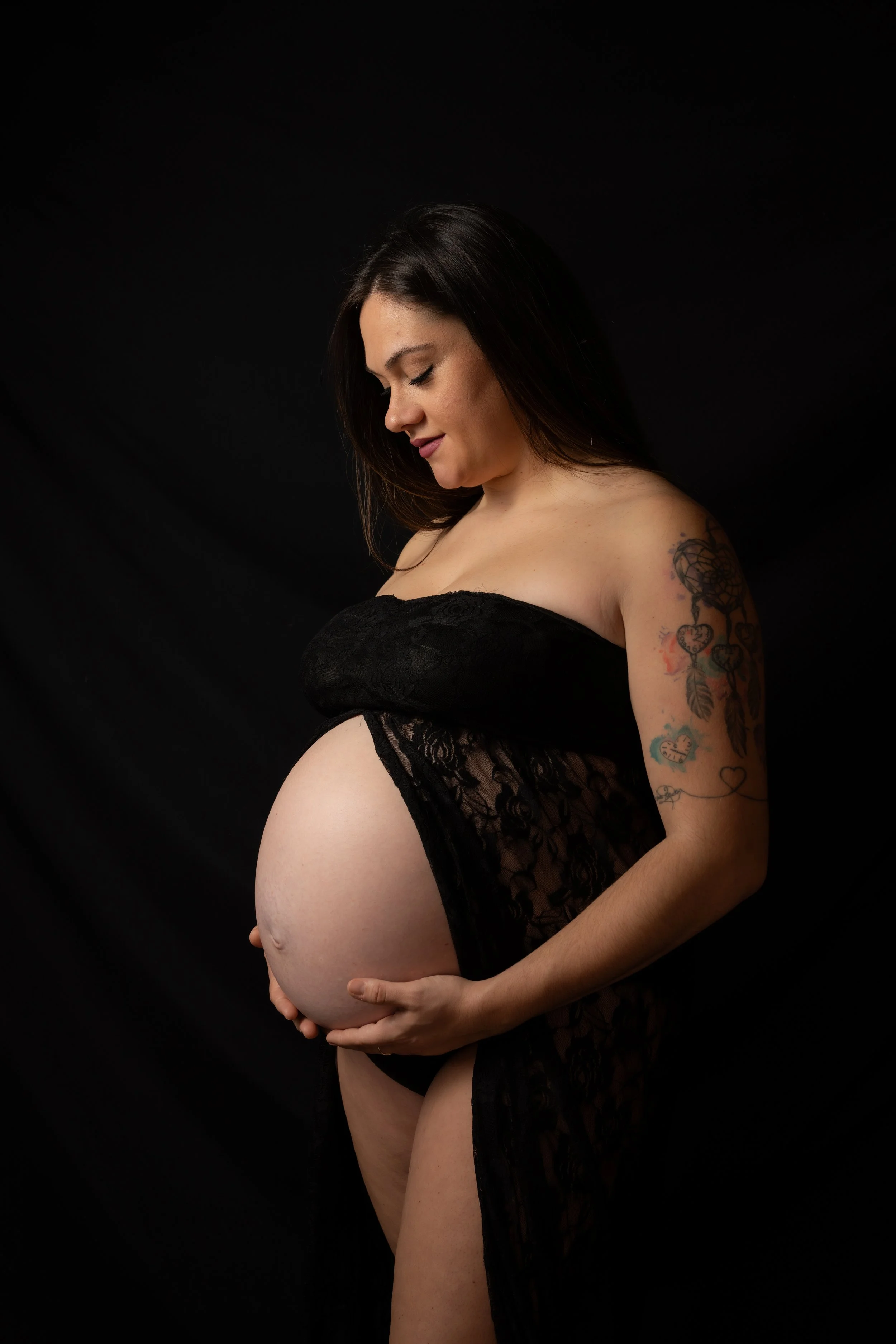 Mujer embarazada con tatuajes sosteniendo su vientre contra fondo negro