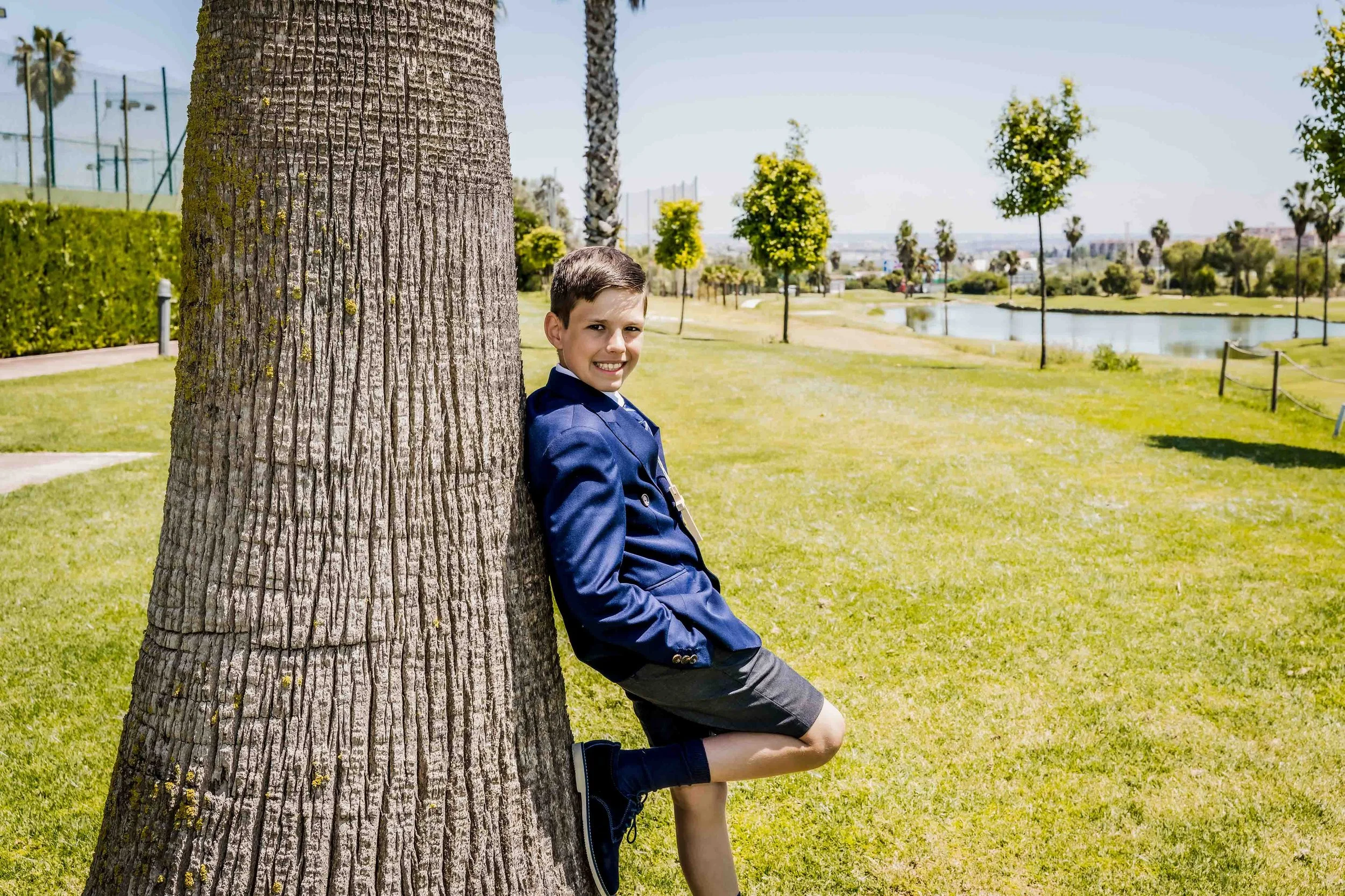 Mi primera comunion, fotografo de comuniones en Sevilla. Niño sonriendo apoyado contra un árbol en un parque con césped, árboles pequeños, un lago y un cielo despejado.