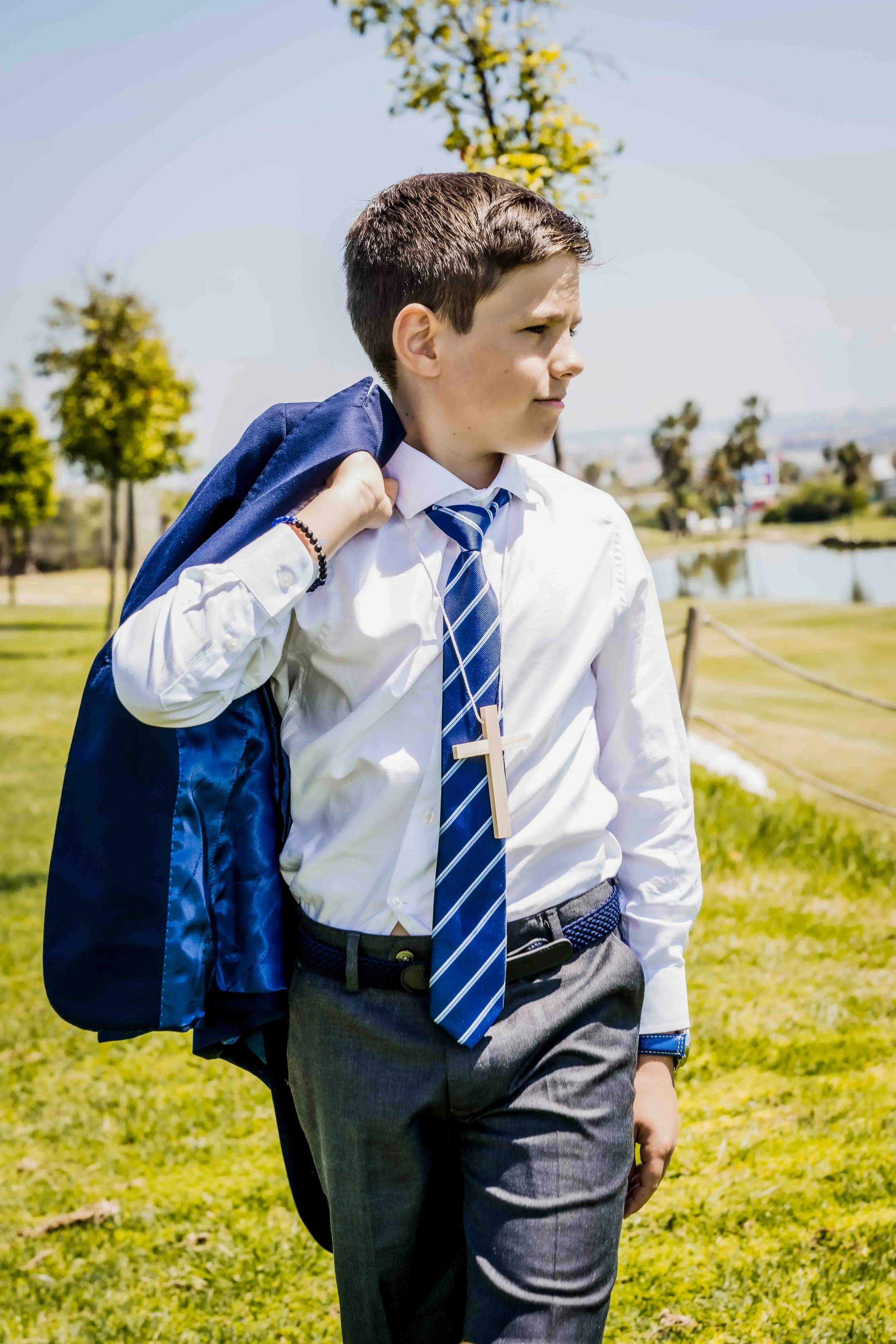 Comuniones en Sevilla, exclusivas, elegantes, diferentes. Fotografo de comuniones. Joven en uniforme escolar con una cruz de madera colgada en el cuello, llevando un saco azul en el hombro, en un parque con árboles y un lago.