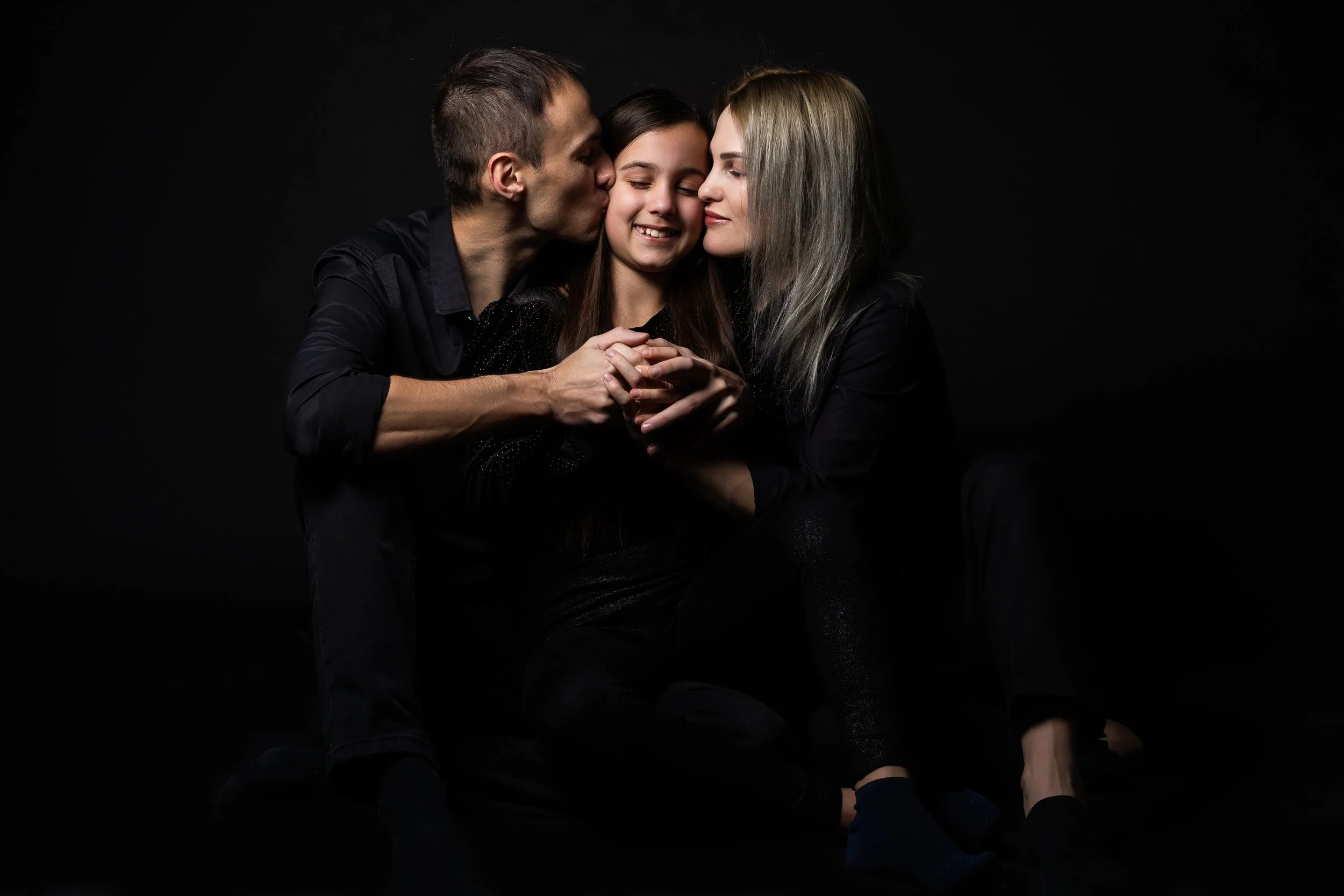 Tres personas en un fondo negro, un hombre besando a una mujer de cabello oscuro, mientras una joven con cabello castaño y cabello rubio con raíces negras mira y sonríe.