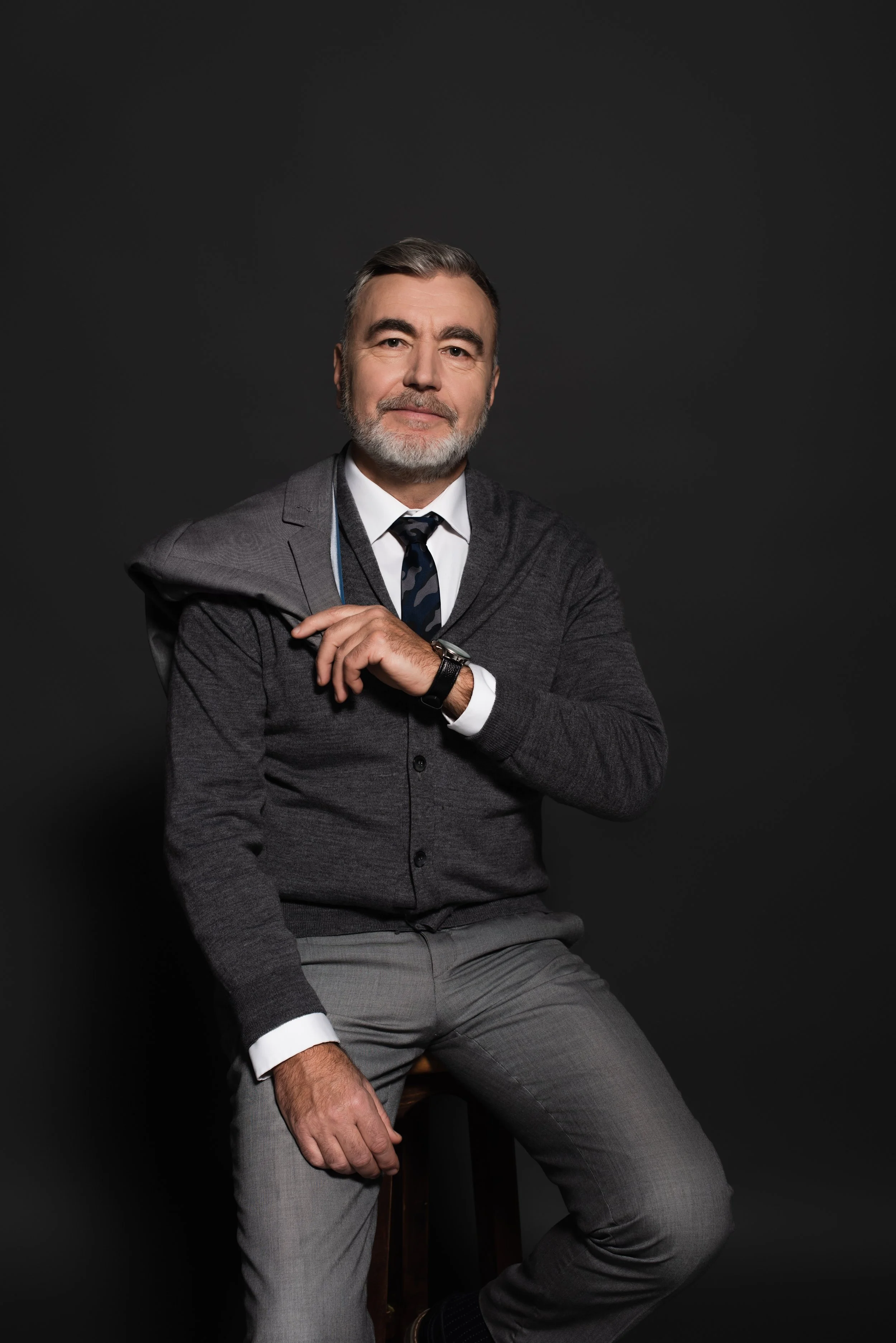 Hombre de cabello gris y barba, con traje sutil y sentirse confiado, sentado en un taburete, en un fondo oscuro.